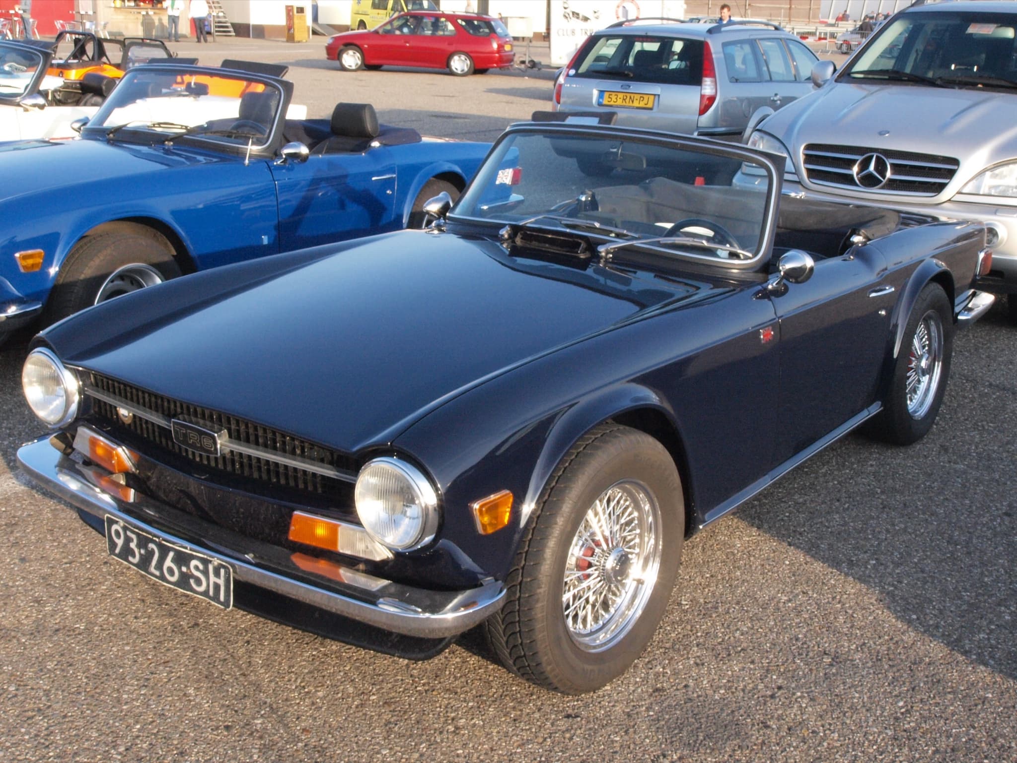A dark blue Triumph TR6 with chrome wire wheels and the hood folded, parked in a paddock car park. Opens a larger view