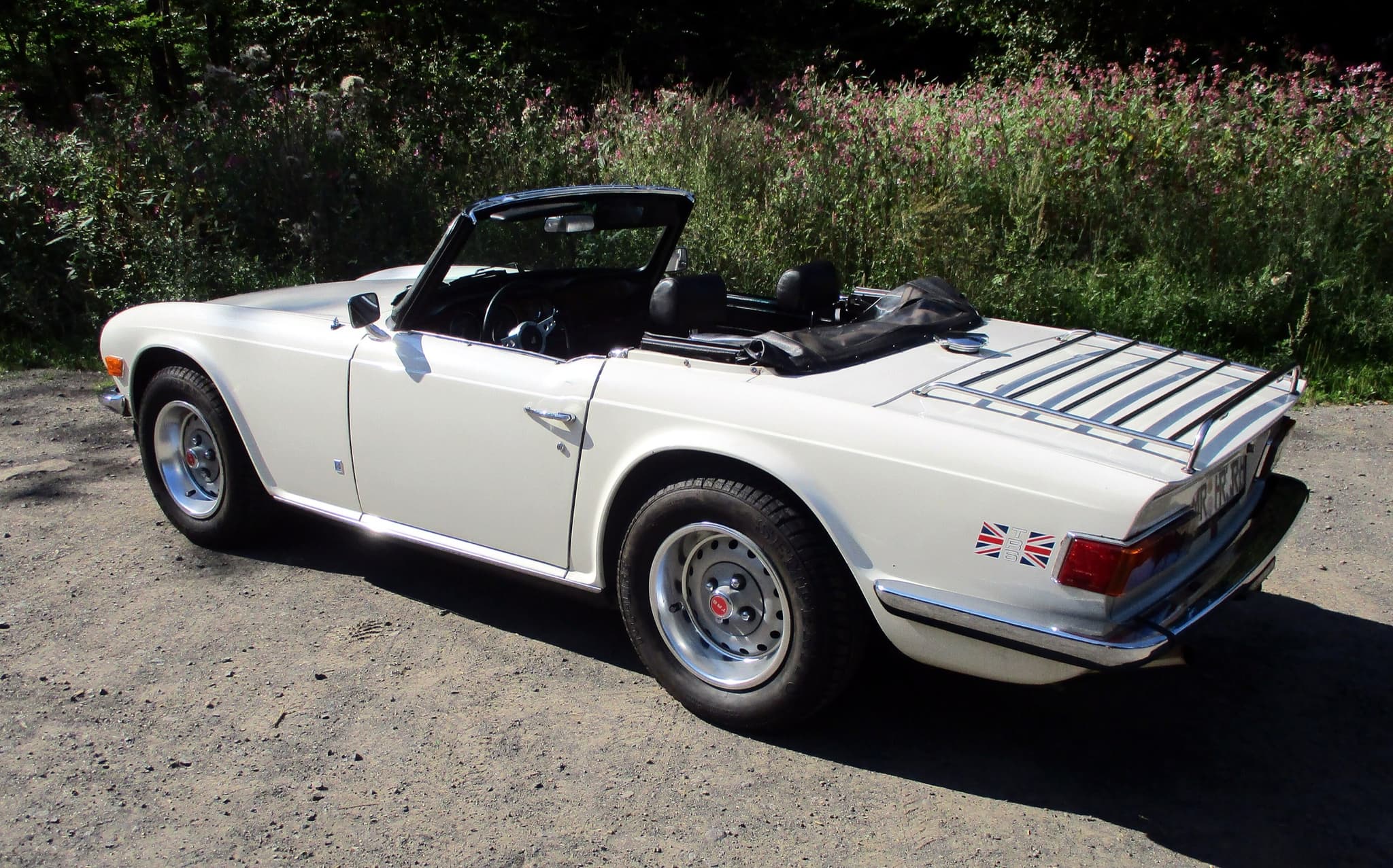 A white Triumph TR6 with a luggage rack, seen from behind on a gravel forest track in strong sun. Opens a larger view