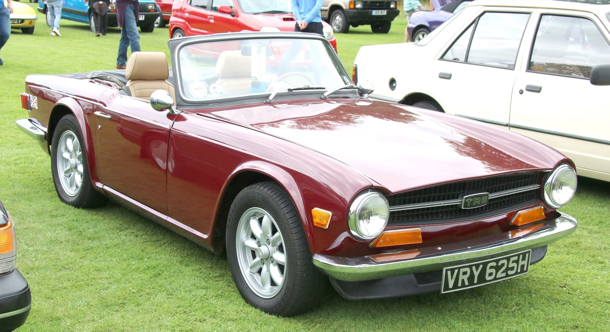 A maroon Triumph TR6 with tan seats, the hood folded and a black front spoiler, on the grass at a car show.