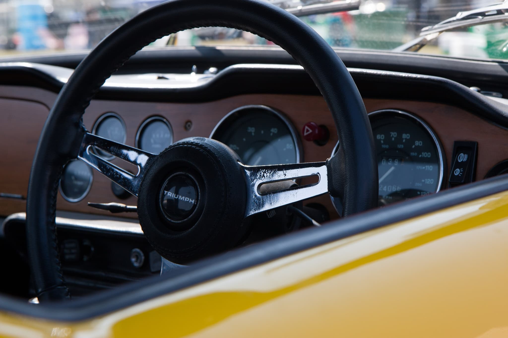 A leather-rimmed steering wheel with slotted chrome spokes in front of the wood facia of a yellow Triumph TR6. Opens a larger view