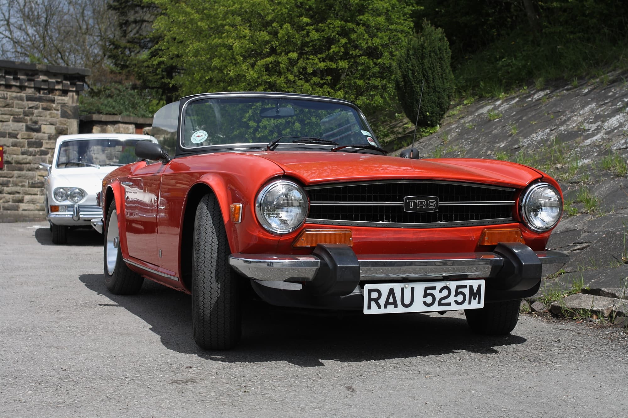 A red Triumph TR6 with black rubber bumper guards and a front spoiler, parked below a stone wall beside a white saloon.