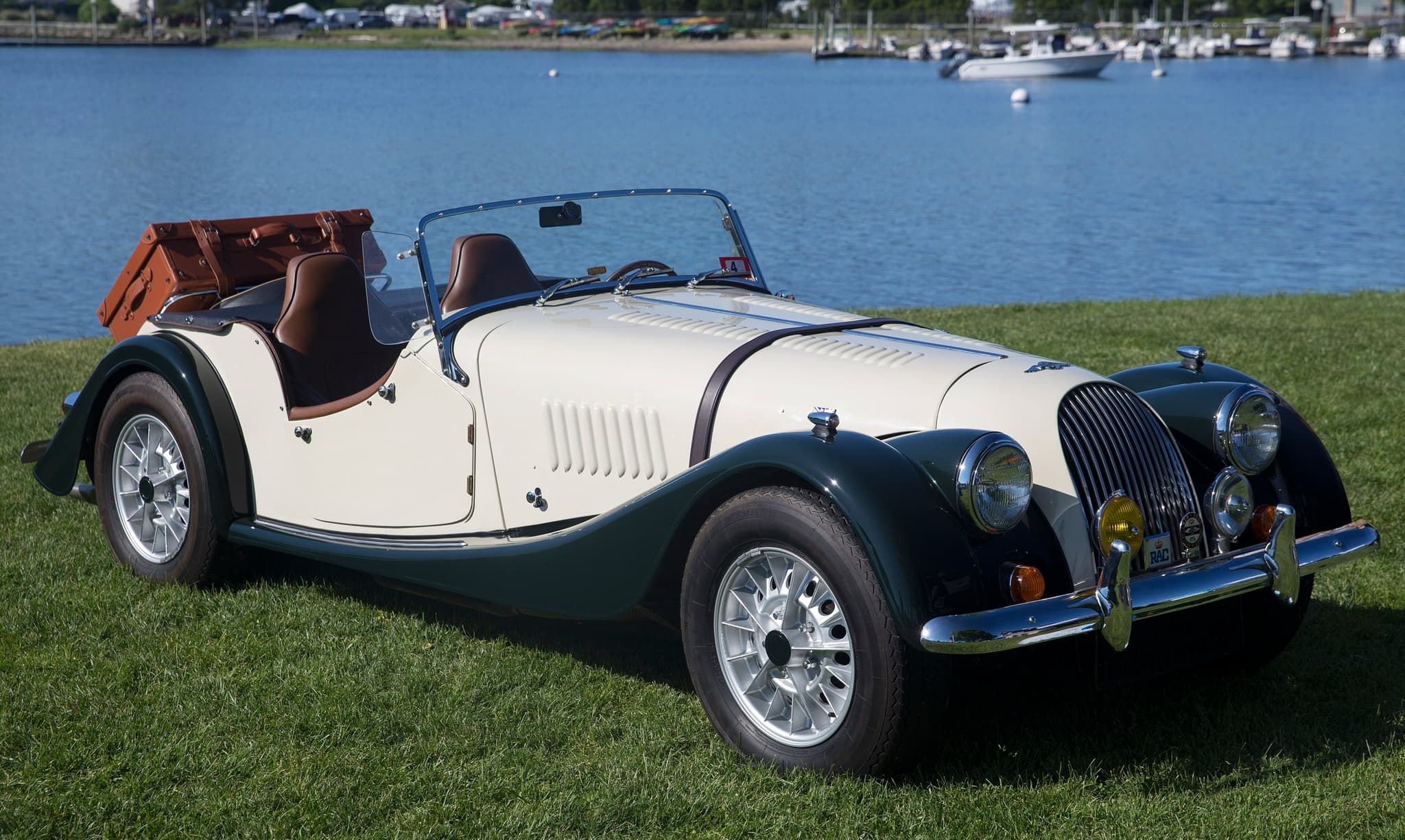 An ivory Morgan Plus 8 with dark green wings and a luggage trunk on the rack, on grass beside a harbour.
