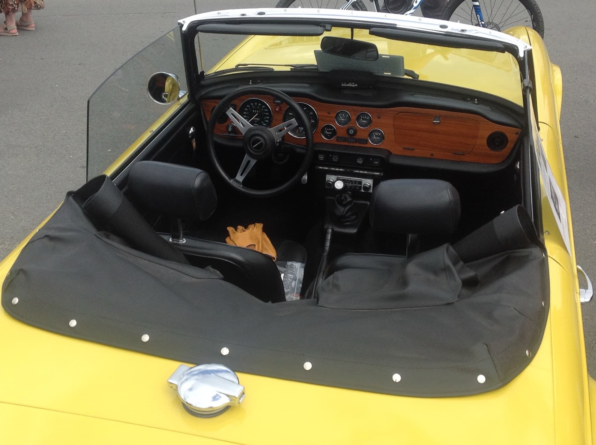 Looking down into a yellow Triumph TR6 from behind: seats with head restraints on stalks, a wood facia, a three-spoke wheel and a tonneau over the passenger side. Opens a larger view