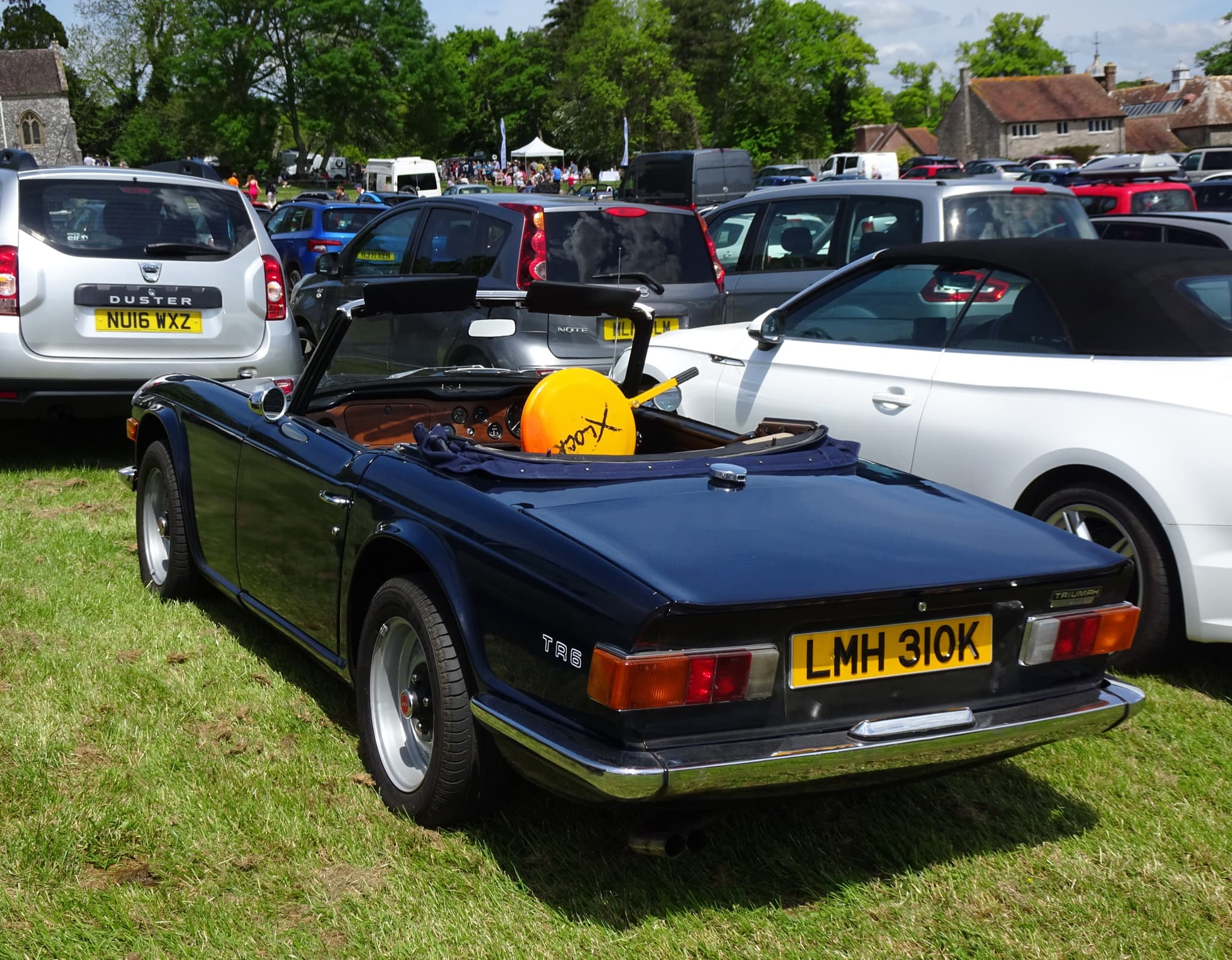 A dark blue right-hand-drive Triumph TR6 seen from behind on the grass of a car park, a church and village houses beyond.