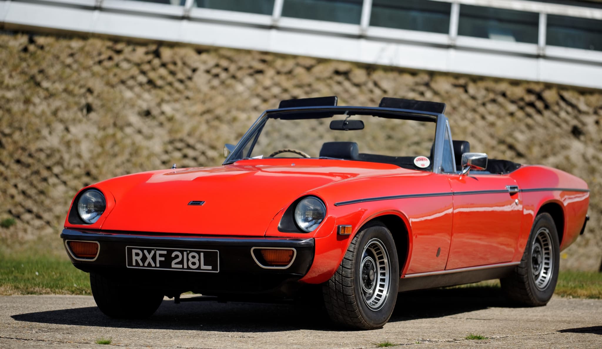 A red Jensen-Healey roadster with the hood folded, on a concrete apron below a grassy bank.