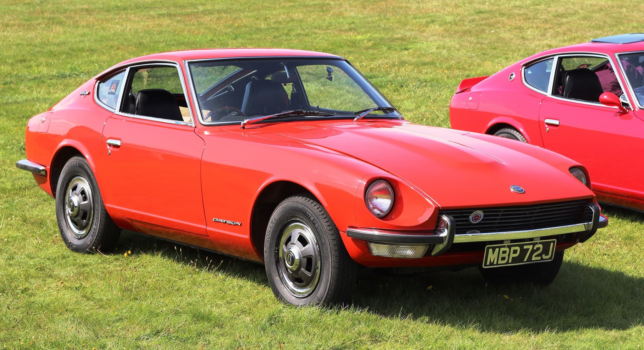 An orange-red Datsun 240Z coupe on mown grass in sunshine.