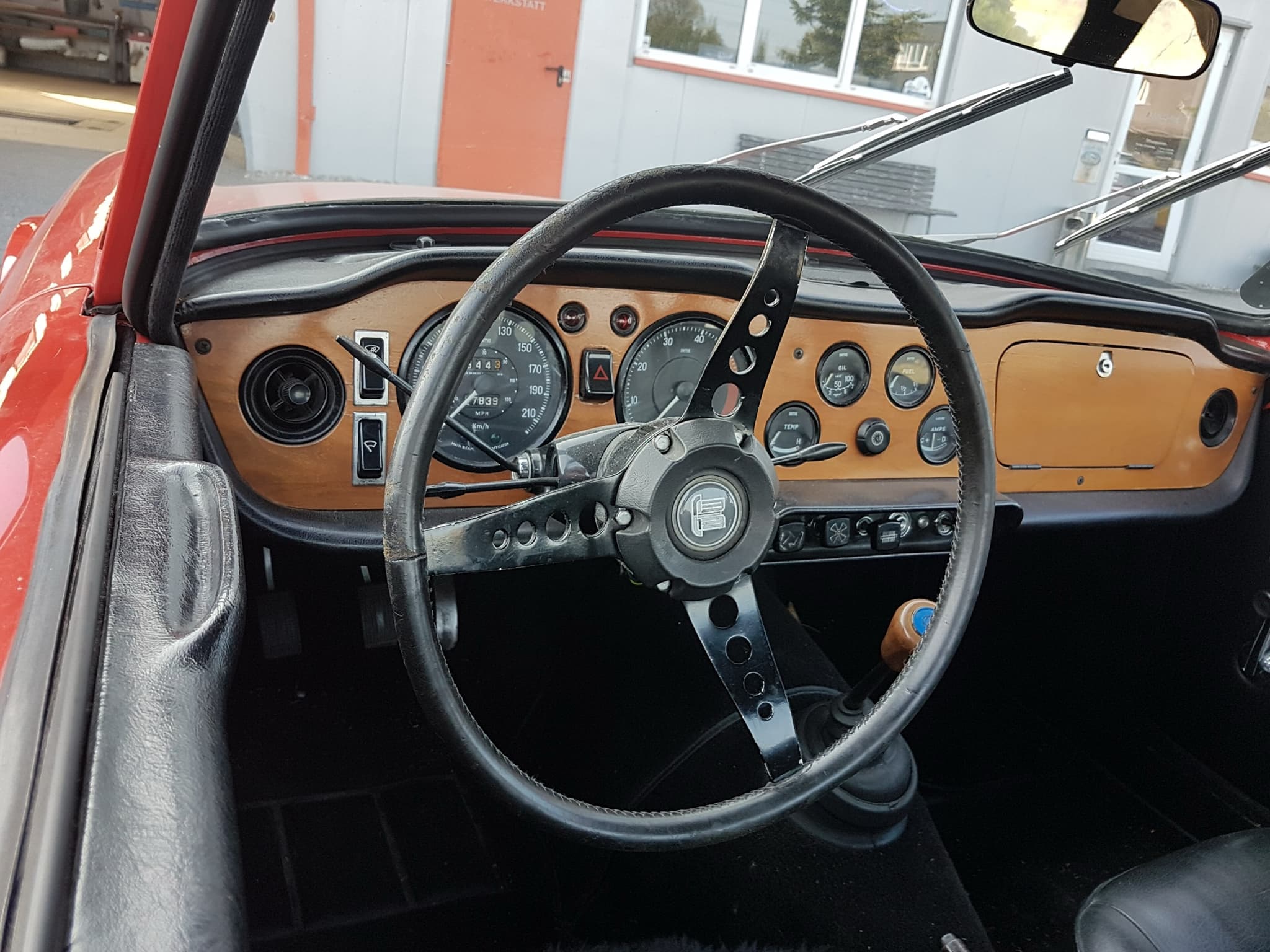 The cockpit of a red left-hand-drive Triumph TR6 seen through the open door: a wood facia with two large dials and a black three-spoke steering wheel. Opens a larger view