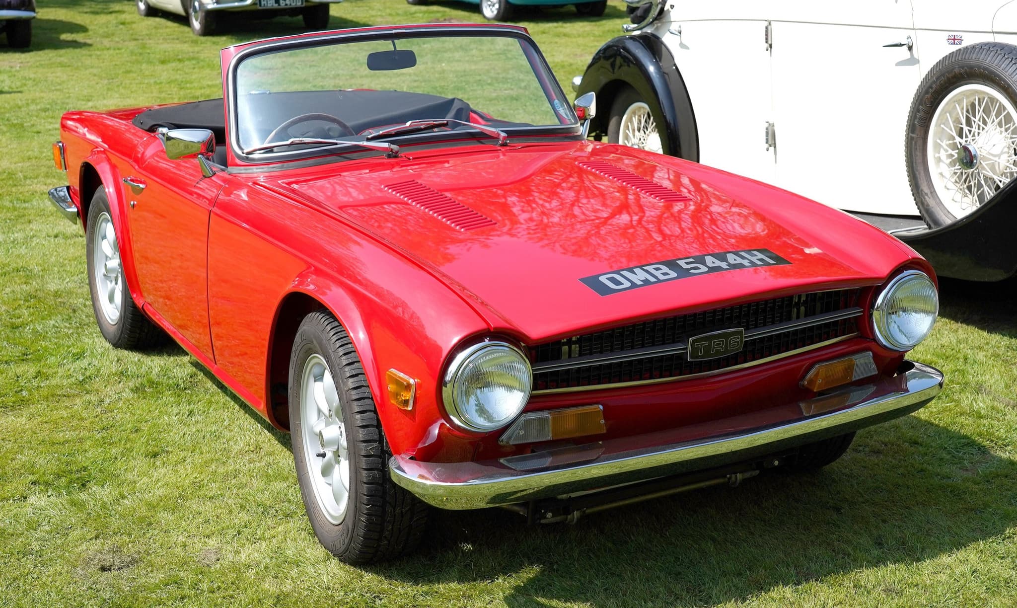 A red Triumph TR6 with two louvred vent panels in its bonnet and alloy wheels, on sunny grass.