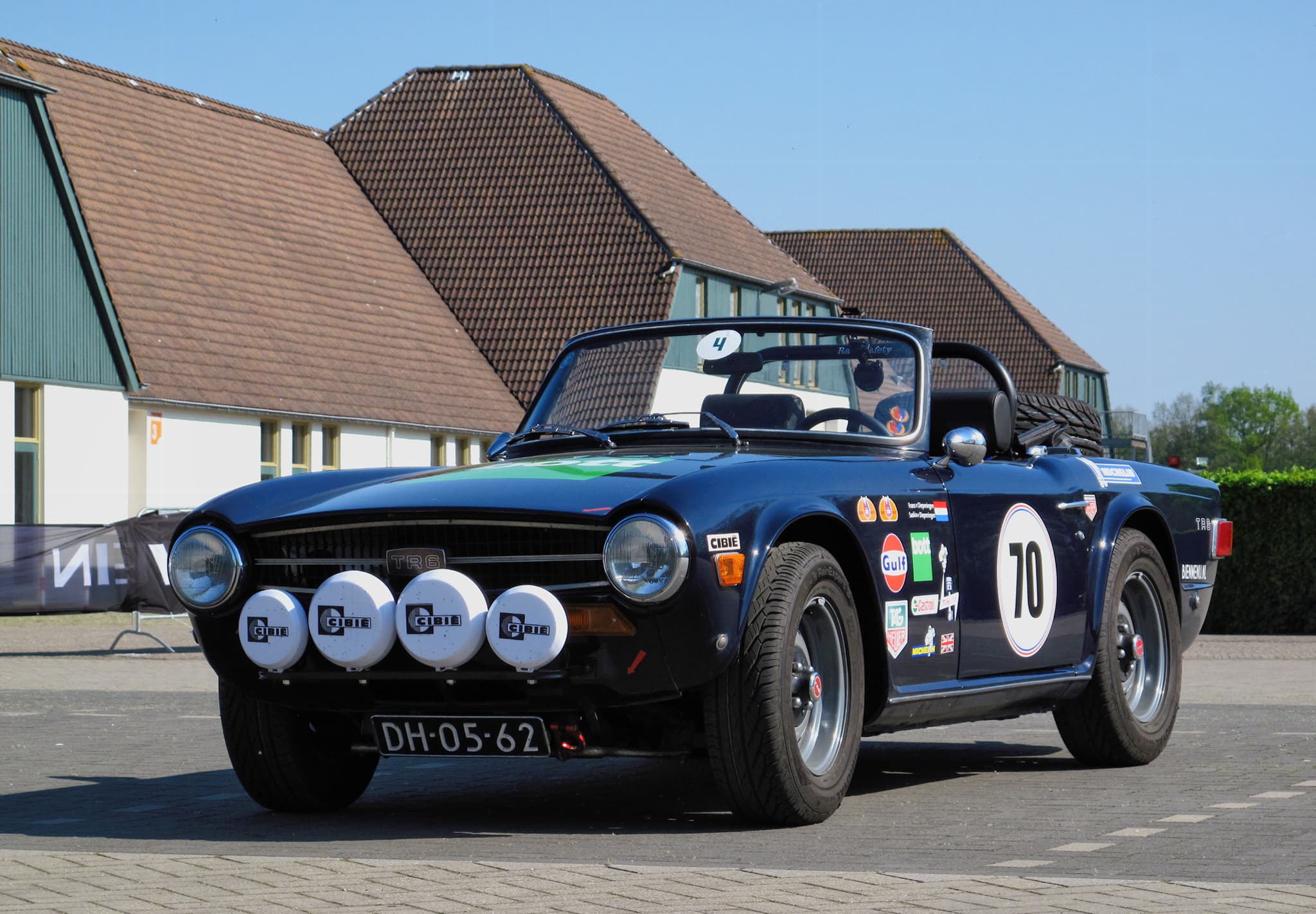 A dark blue Triumph TR6 with four spot lamps, rally plates and the number 70, parked on brick paving under a blue sky.