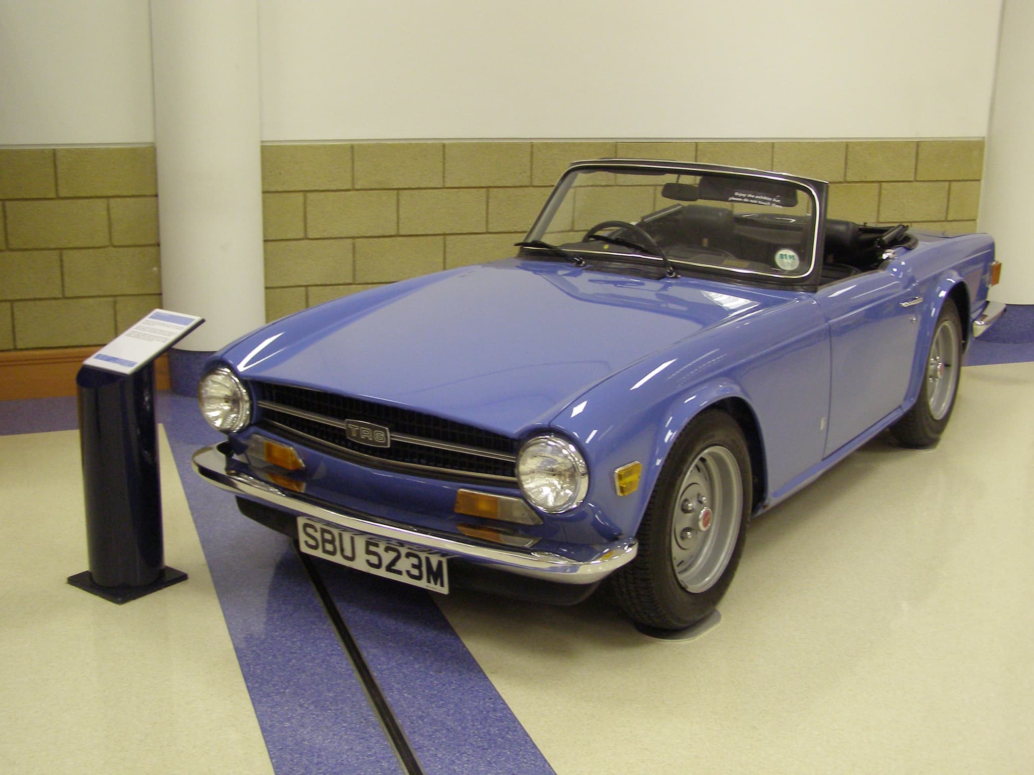 A mid-blue Triumph TR6 with a black front spoiler on a cream museum floor. Opens a larger view