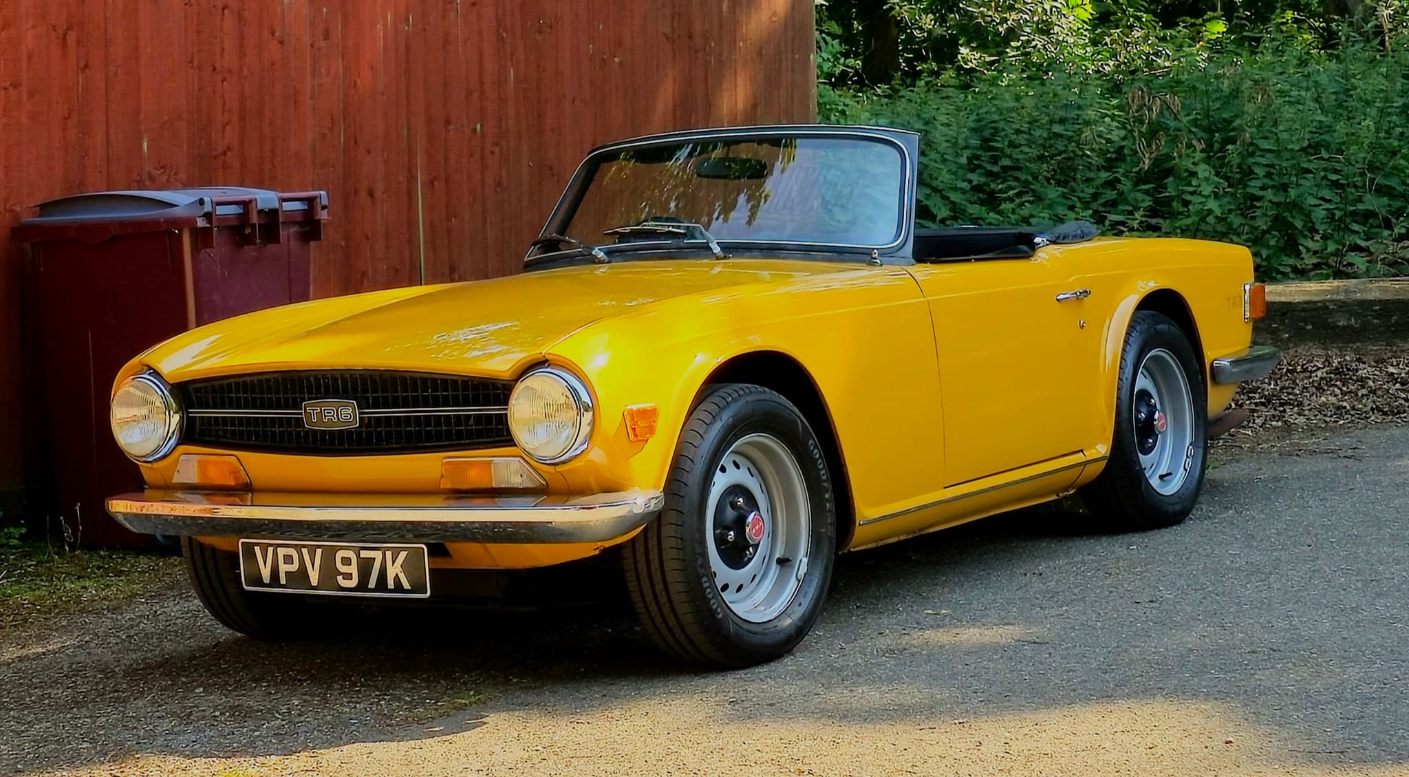 A yellow Triumph TR6 with its hood folded, parked on a gravel drive in front of a red timber fence.