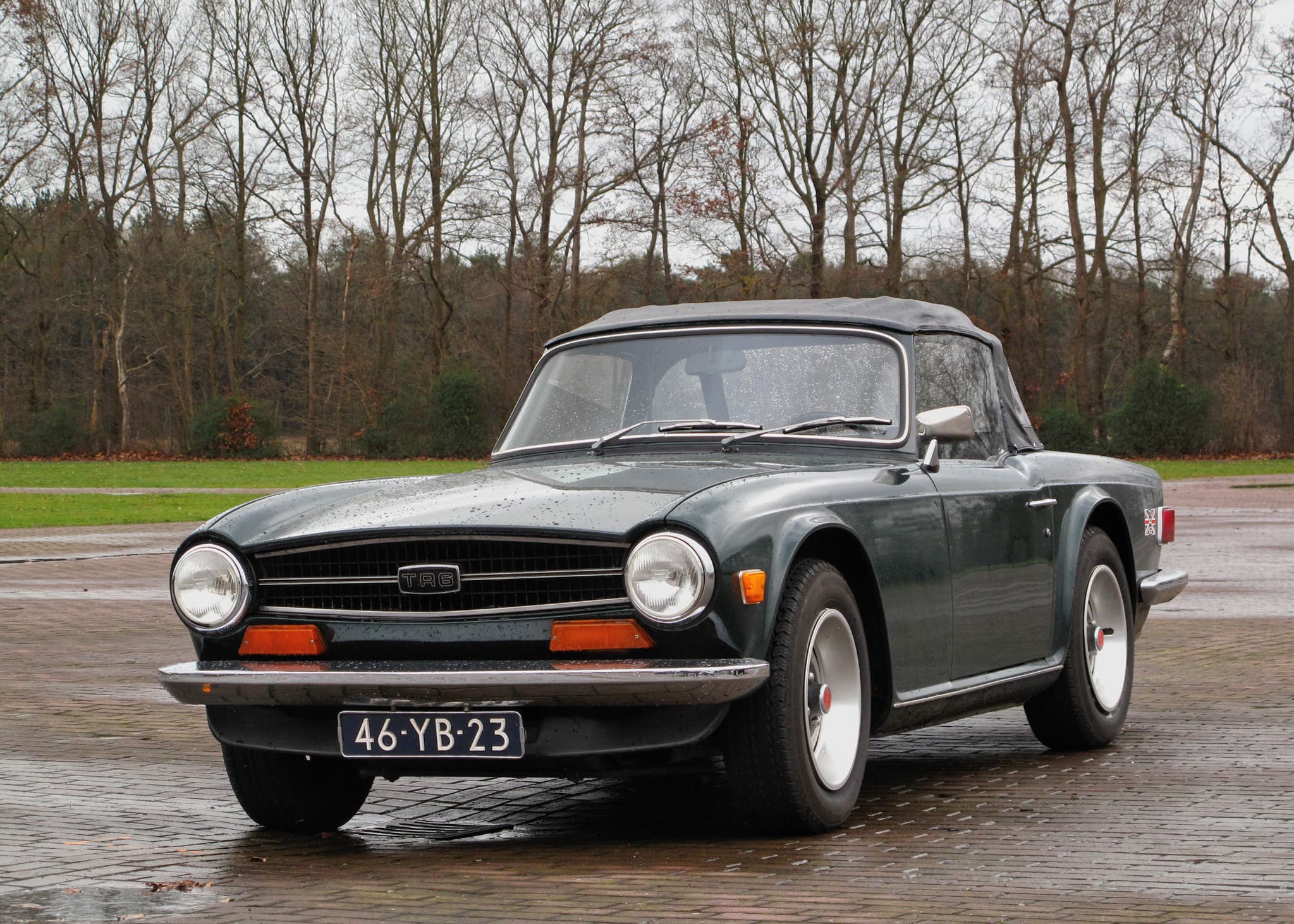 A dark green Triumph TR6 with its grey hood up, parked on wet brick paving under bare winter trees. Opens a larger view