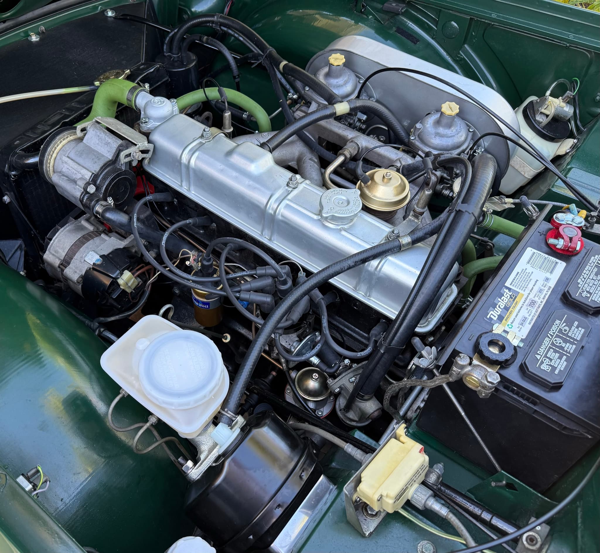 Looking down into a dark green Triumph TR6's engine bay: a six-cylinder engine with twin carburettors, an air pump and emission-control pipework.