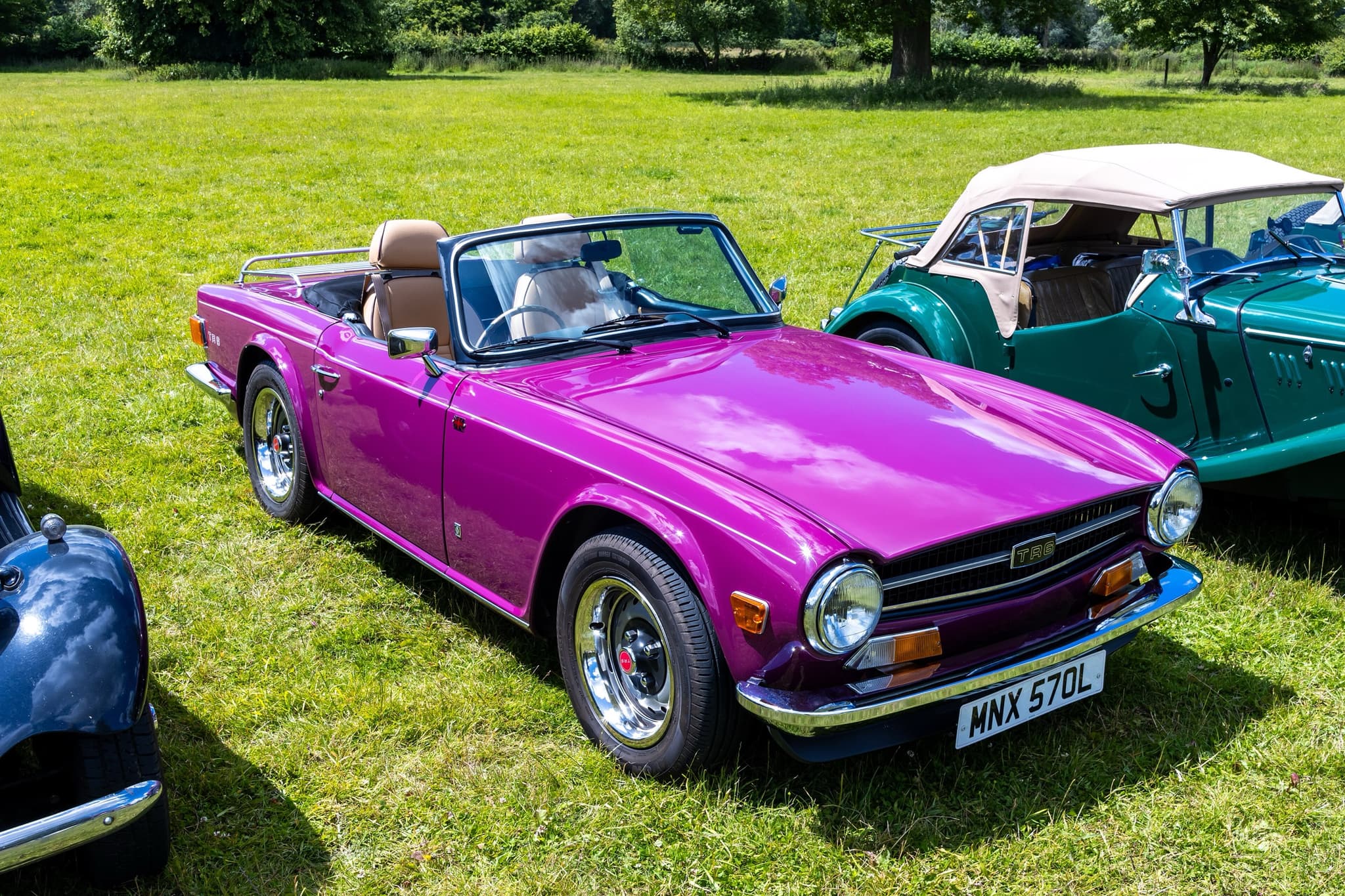 A magenta Triumph TR6 with tan seats, a luggage rack and a black front spoiler, on mown grass beside a green Morgan. Opens a larger view