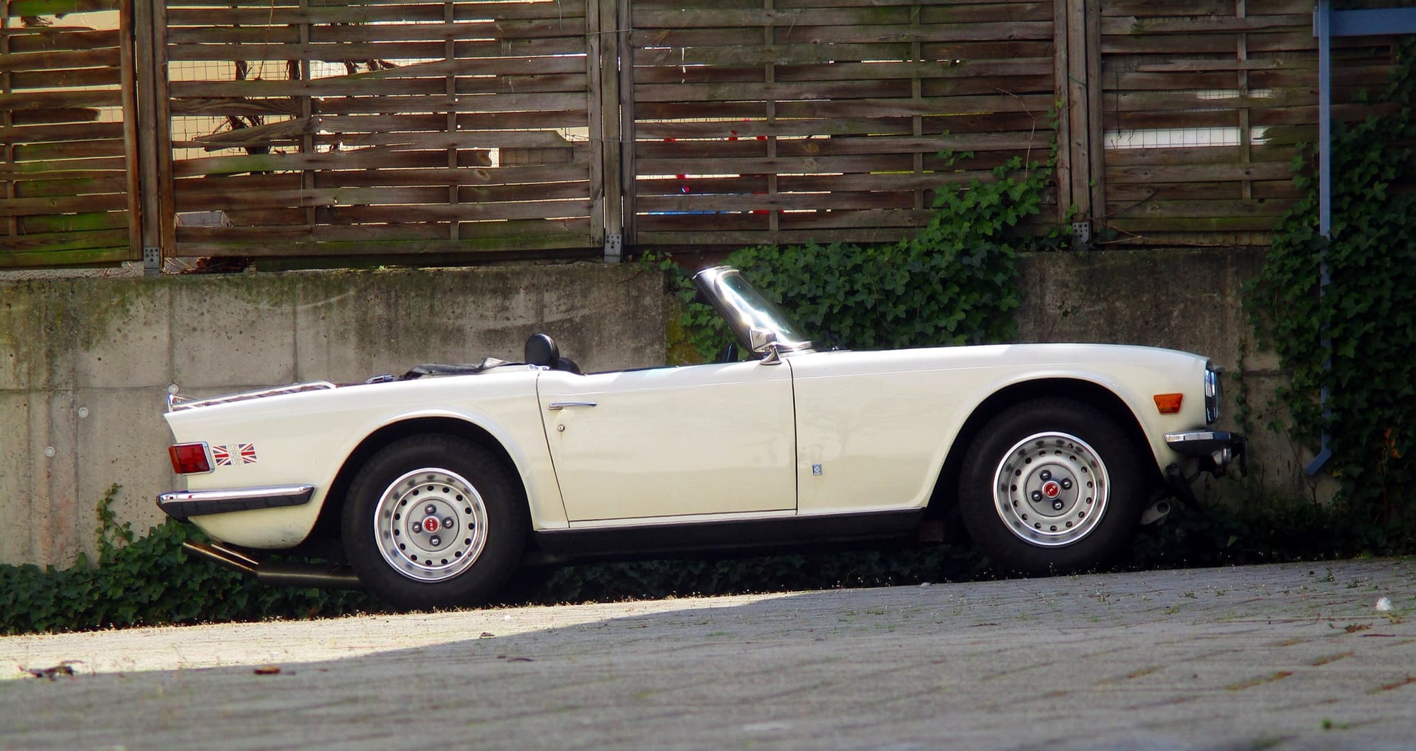 A white Triumph TR6 in side profile with its hood folded, on a paved courtyard before a timber fence.