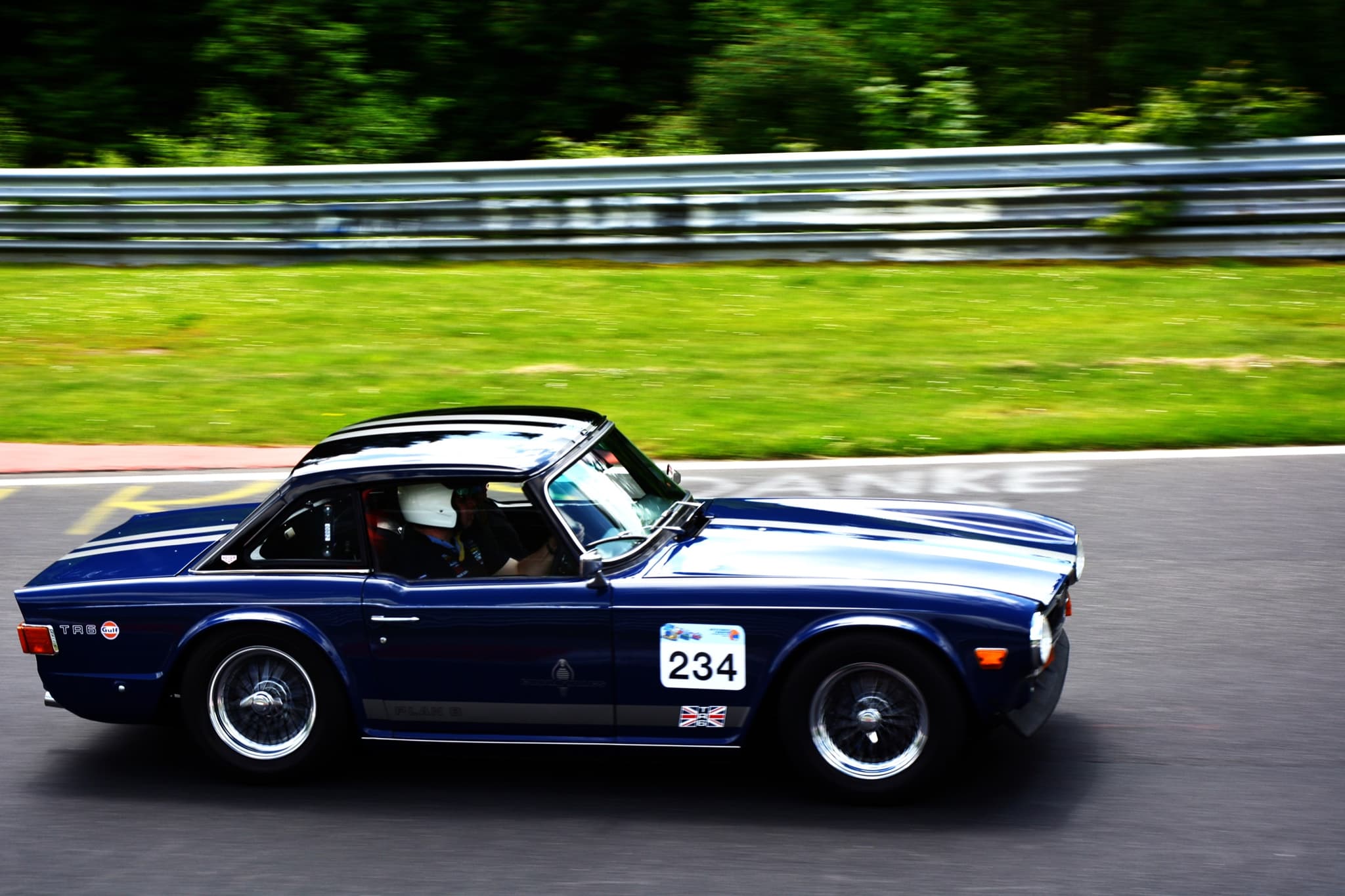 A dark blue Triumph TR6 with a hardtop and white stripes, number 234, at speed on the Nuerburgring.