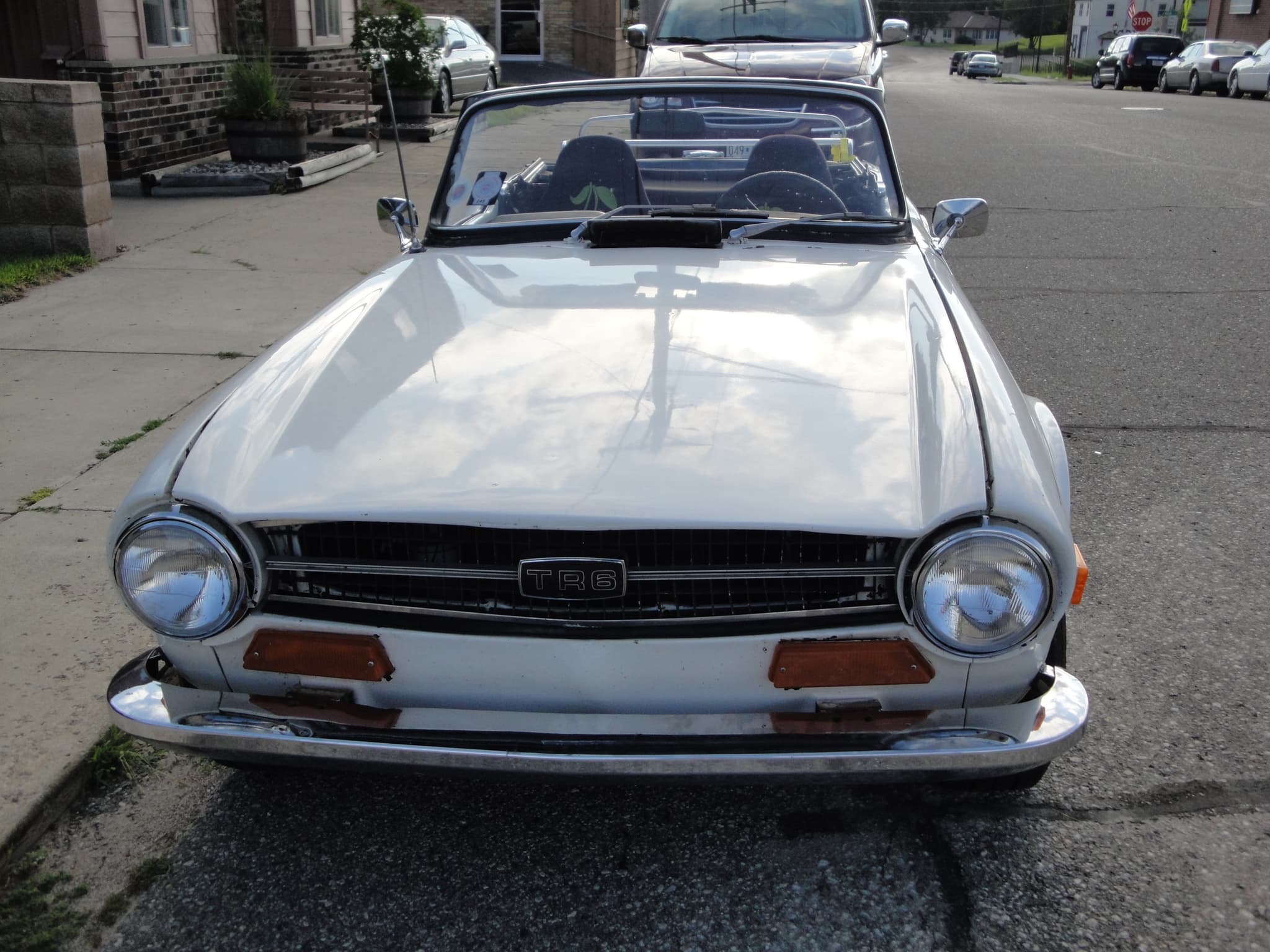 A white Triumph TR6 seen nose-on at a kerb: a full-width grille with the TR6 badge, a headlamp at each end and a plain chrome bumper. Opens a larger view