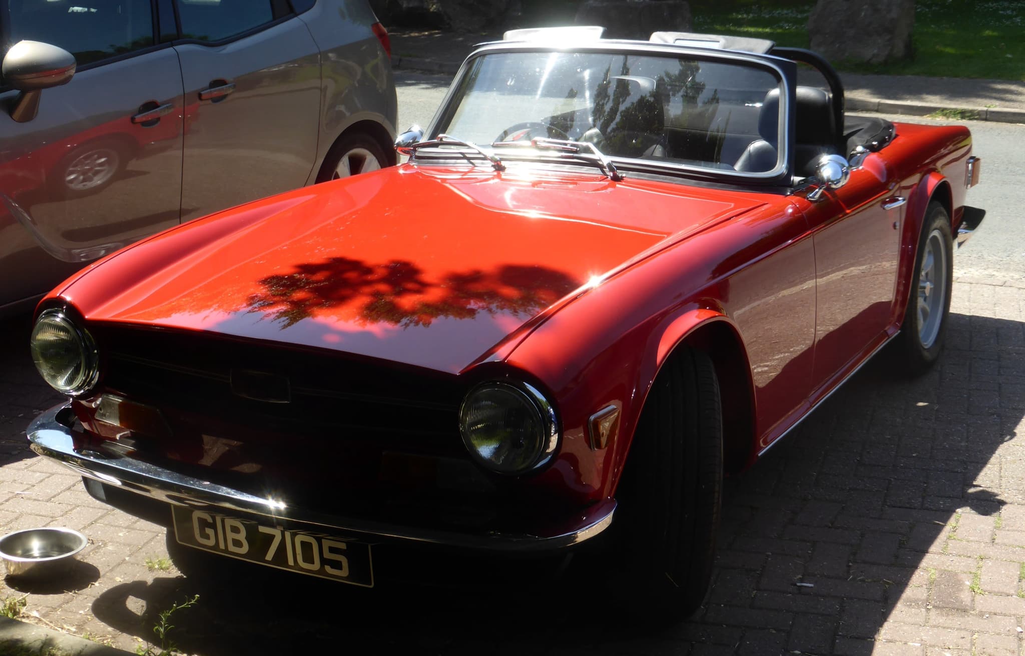 A red right-hand-drive Triumph TR6 with a roll bar, parked on block paving in hard sunlight beside a grey hatchback.