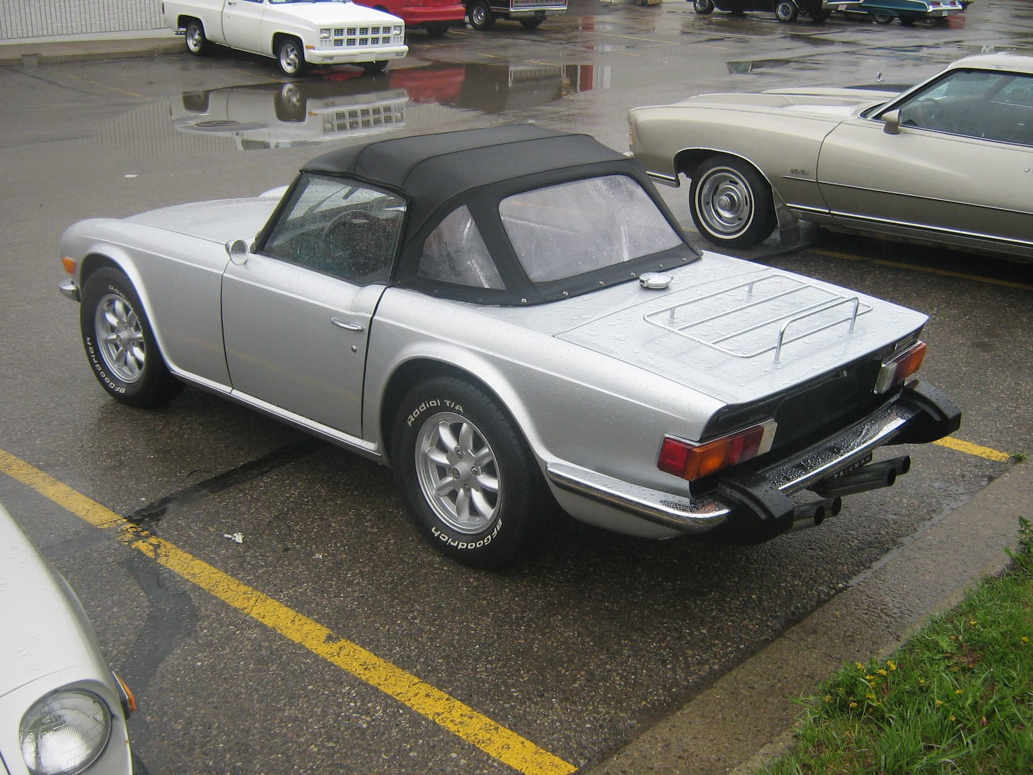 A silver Triumph TR6 with its hood up and black rubber over-riders on the rear bumper, in a wet car park.