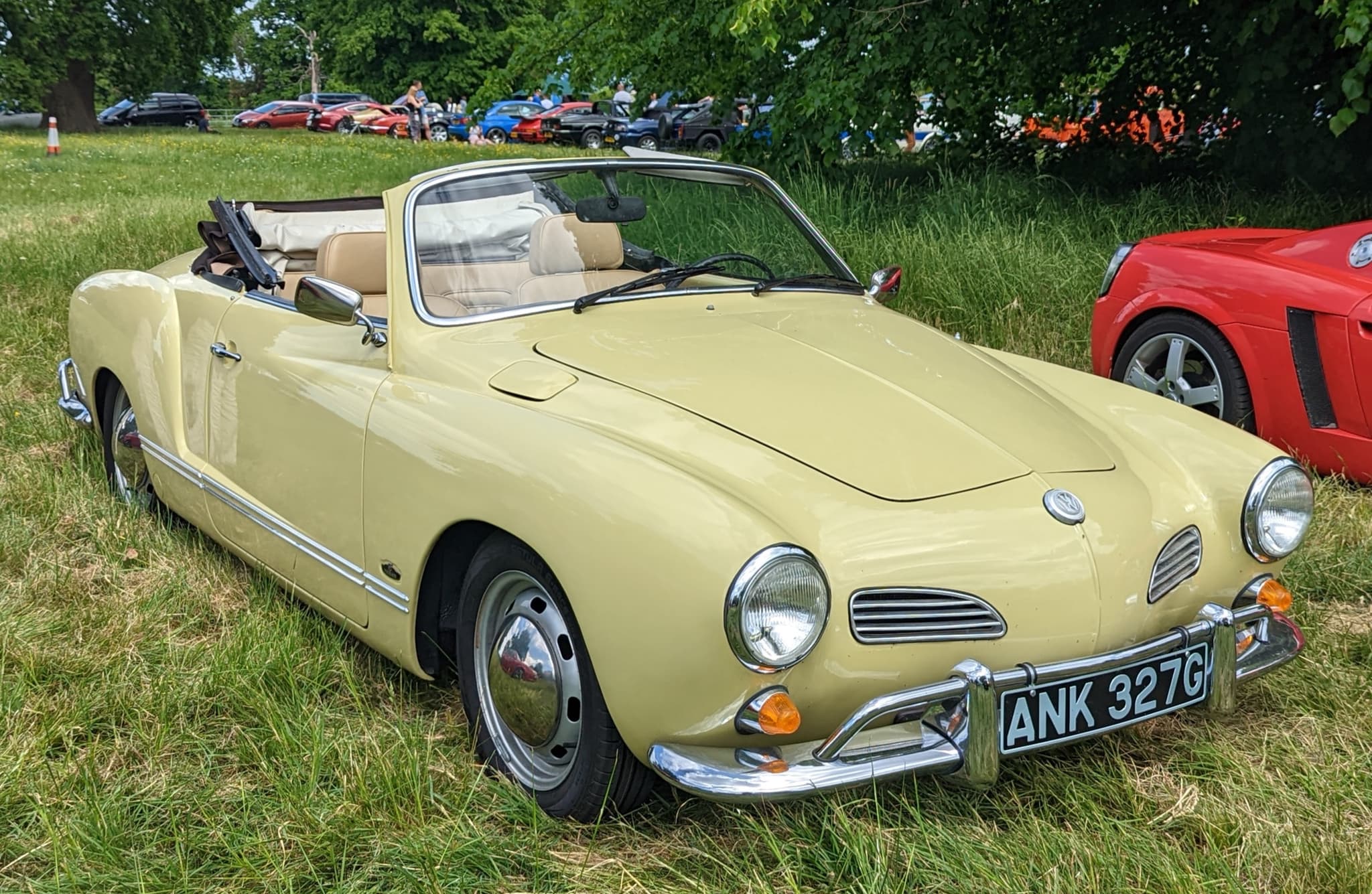 A pale yellow Volkswagen Karmann Ghia convertible with tan seats and the hood folded, on long grass at a car show.
