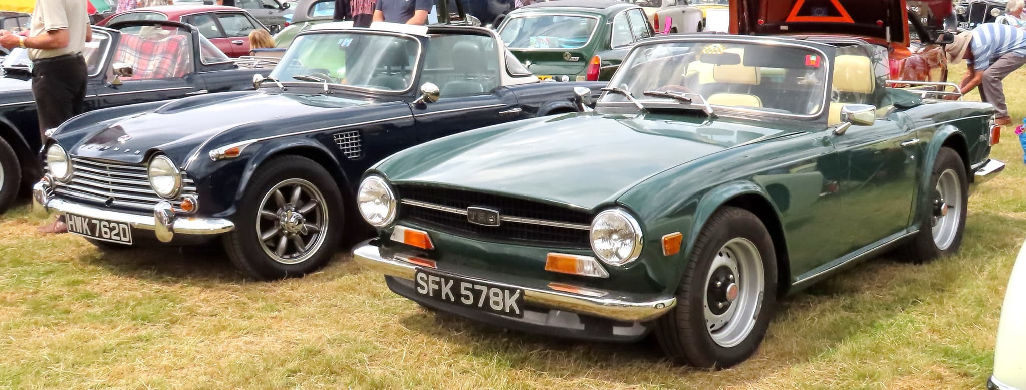 A dark blue Triumph with the older body, its headlamps in the wings, parked beside a dark green TR6 with its headlamps at the ends of a full-width grille.