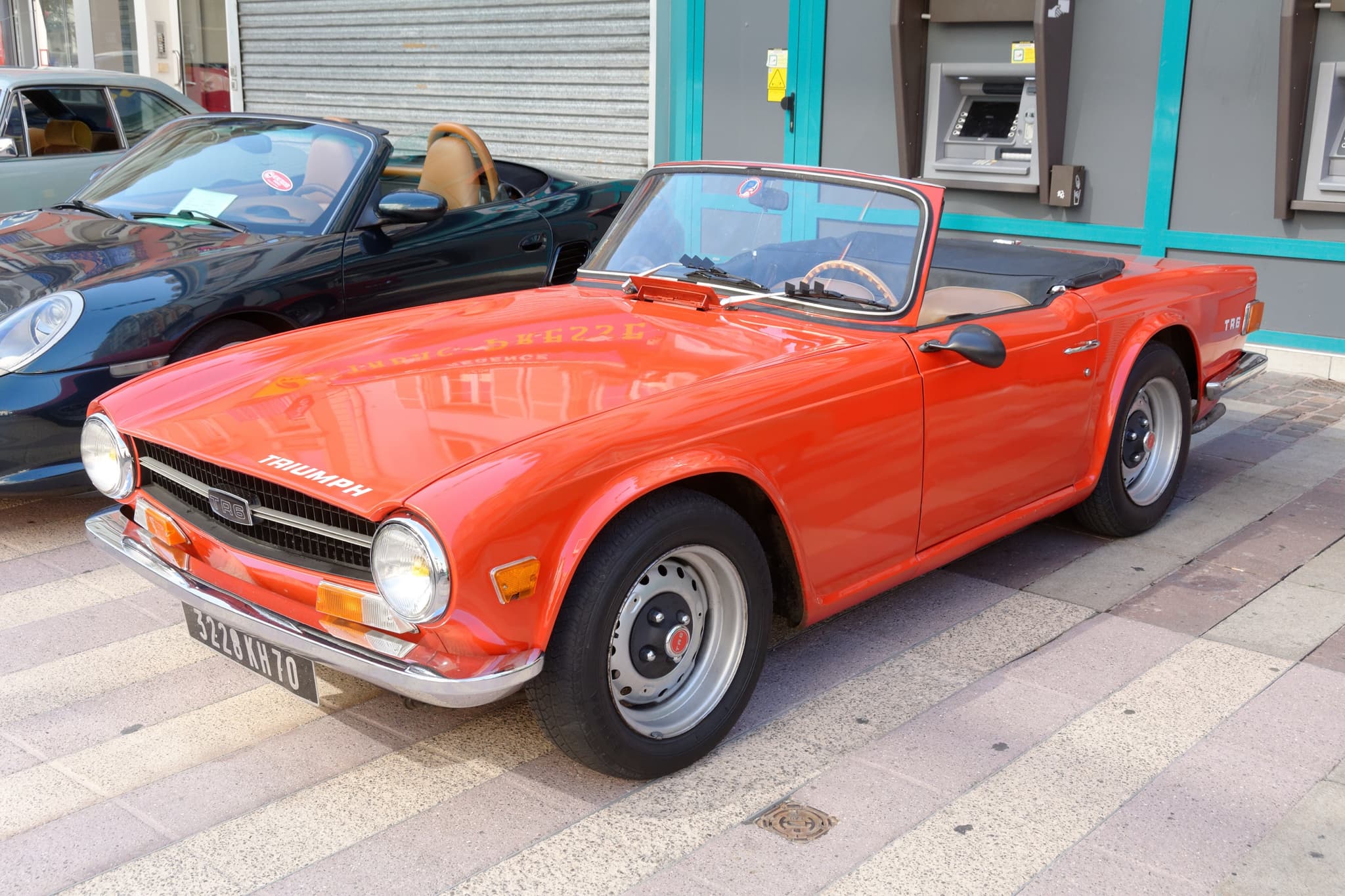 An orange Triumph TR6 with tan seats and a wood-rim steering wheel, parked on pale paving in front of a shopfront. Opens a larger view