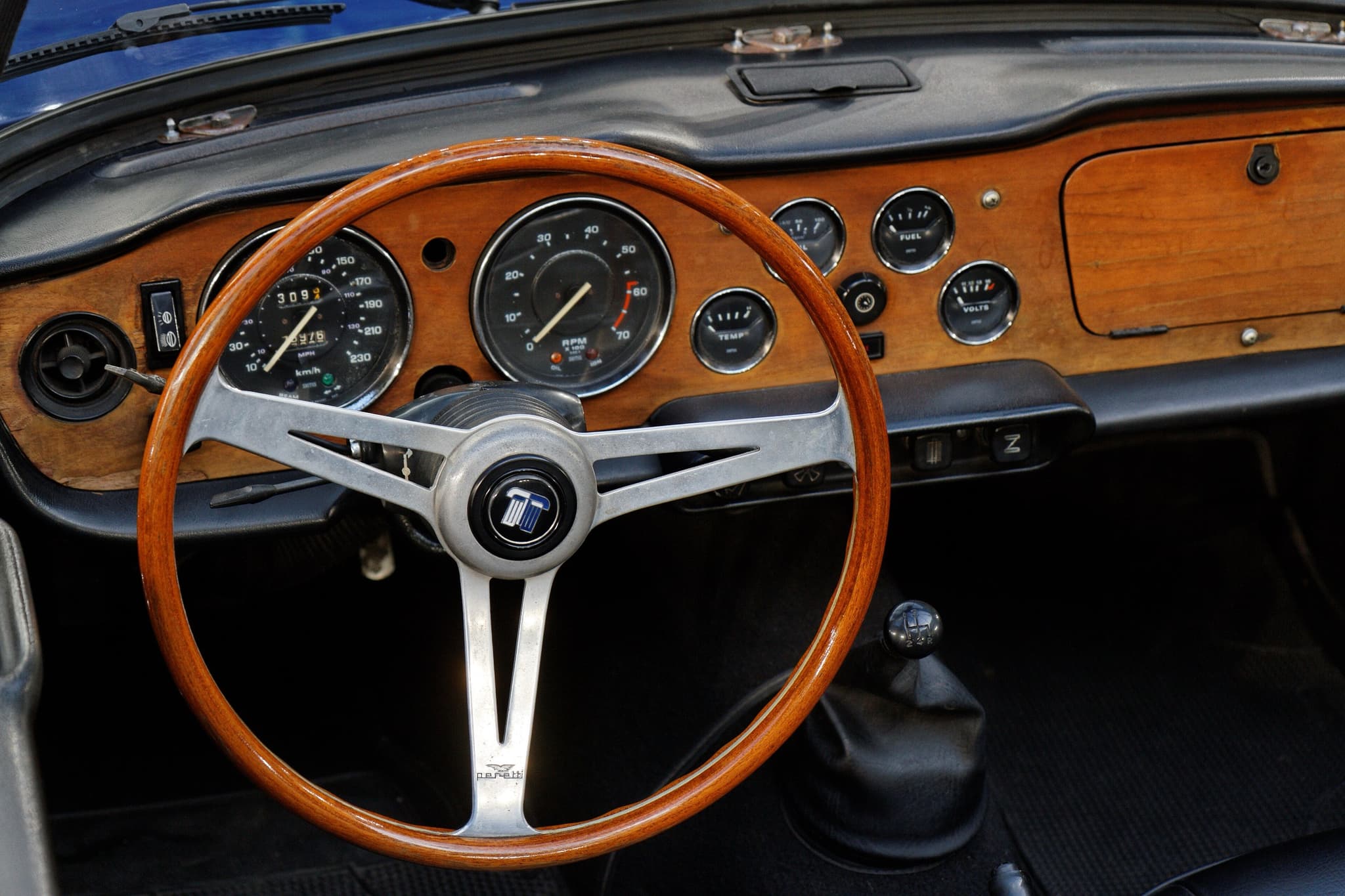 The wood facia of a left-hand-drive Triumph TR6 with its speedometer, tachometer and small gauges, and a wood-rimmed steering wheel.