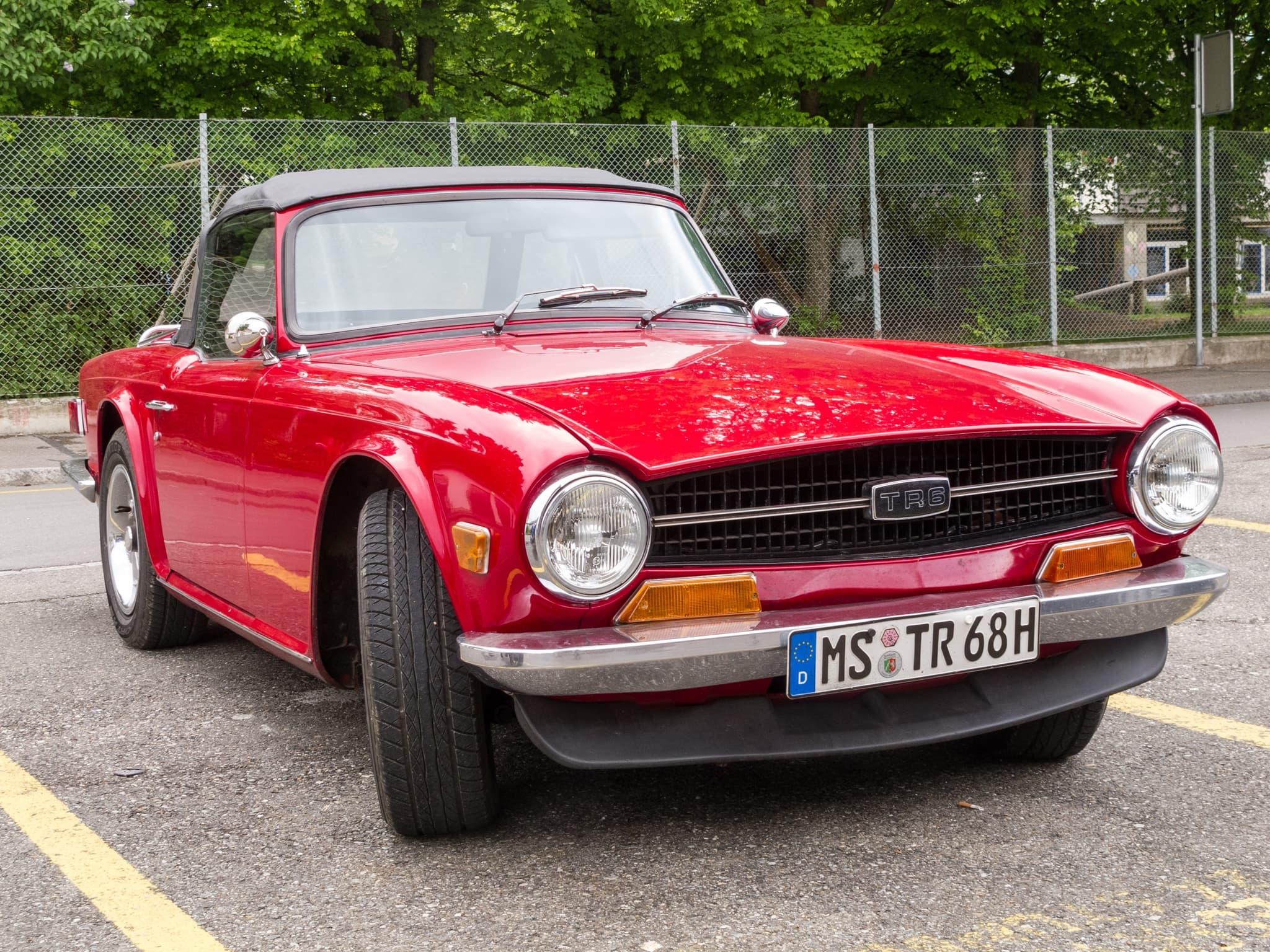 A red Triumph TR6 with its black hood up and a black front spoiler, parked alone in an empty car park.