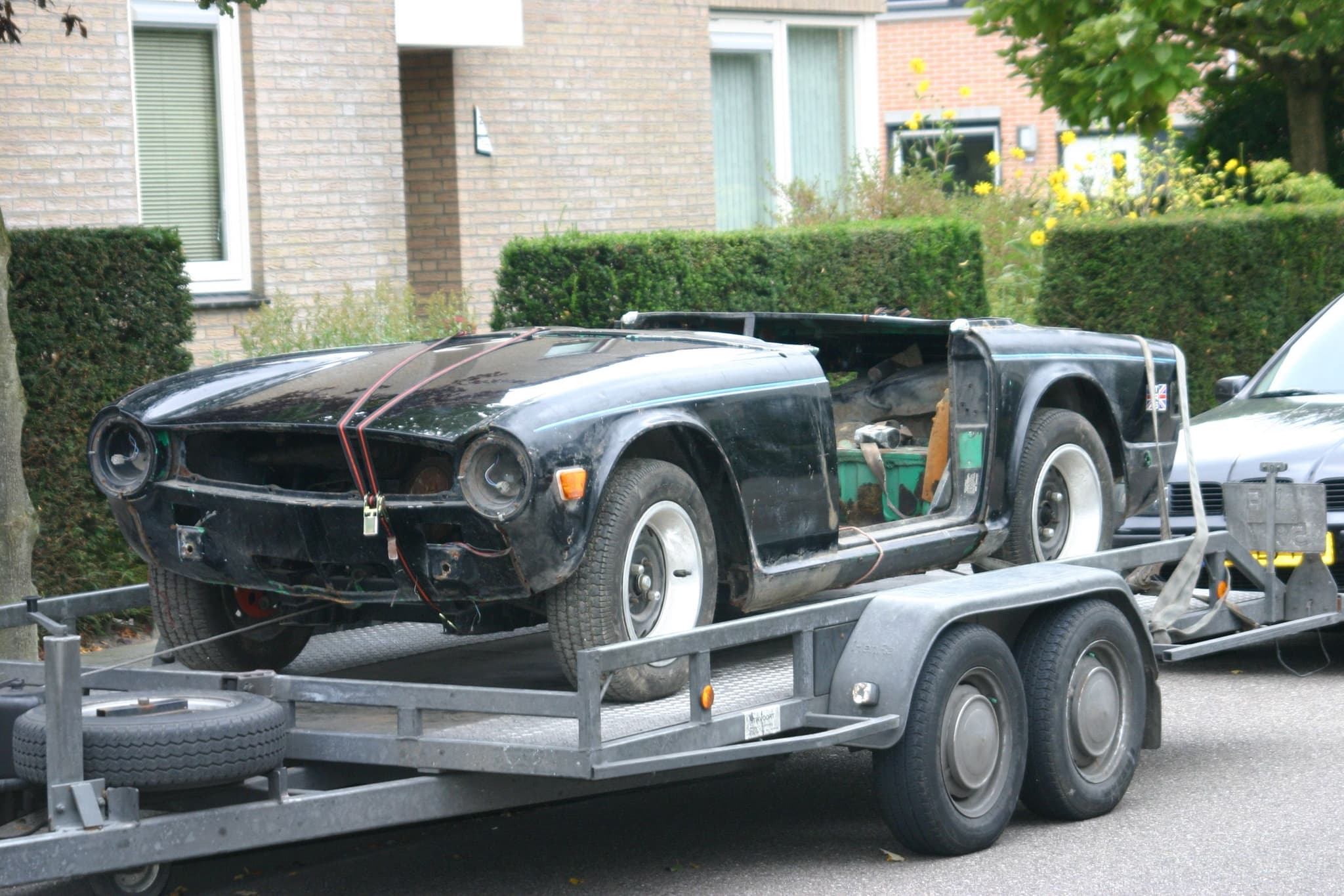 A stripped black Triumph TR6 body on its frame, without bumpers, grille or lamps, strapped to a car trailer in a street.