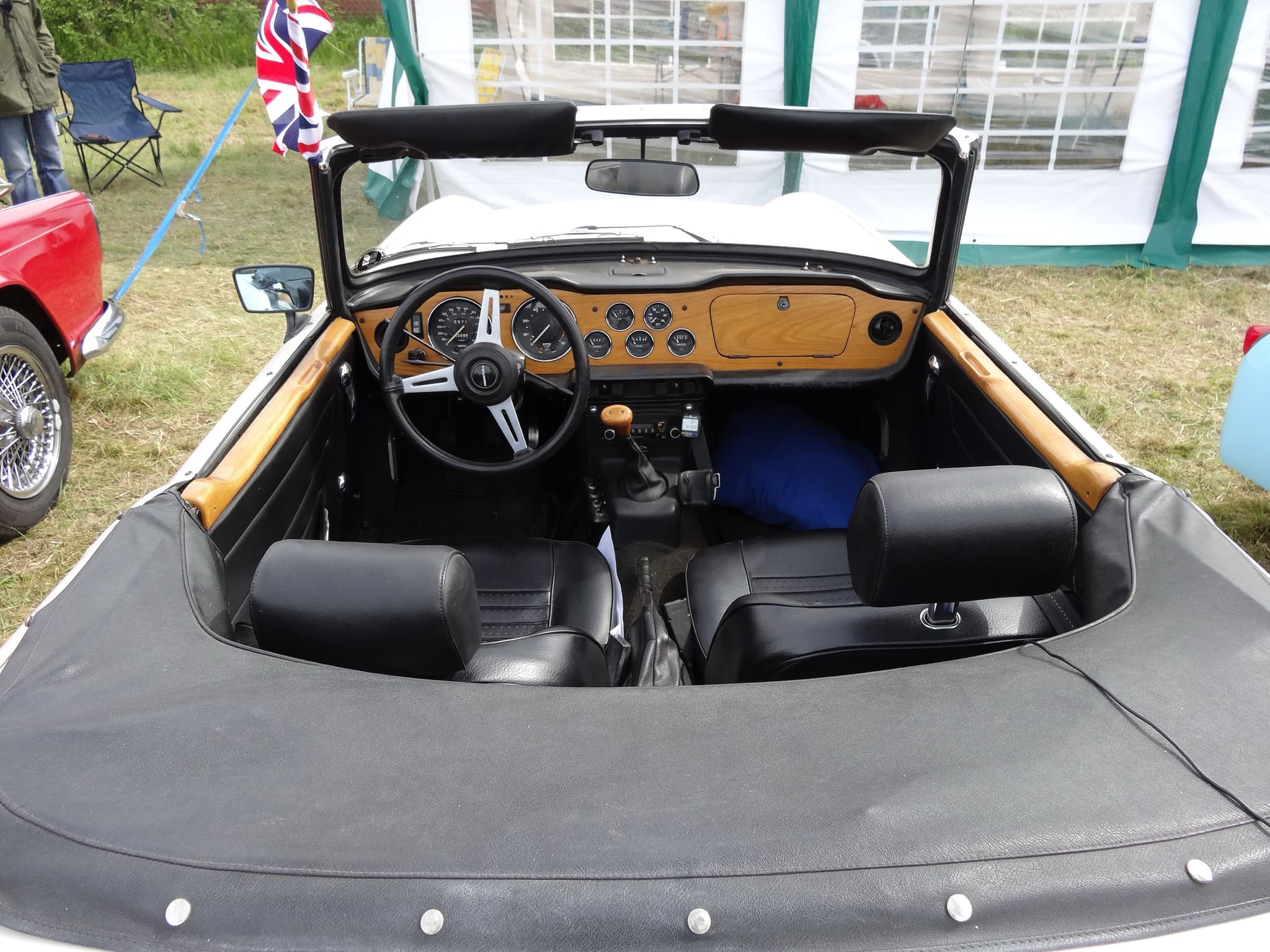 Looking into a white Triumph TR6 from behind: seats with head restraints on stalks, a wood facia and a tonneau over the rear. Opens a larger view