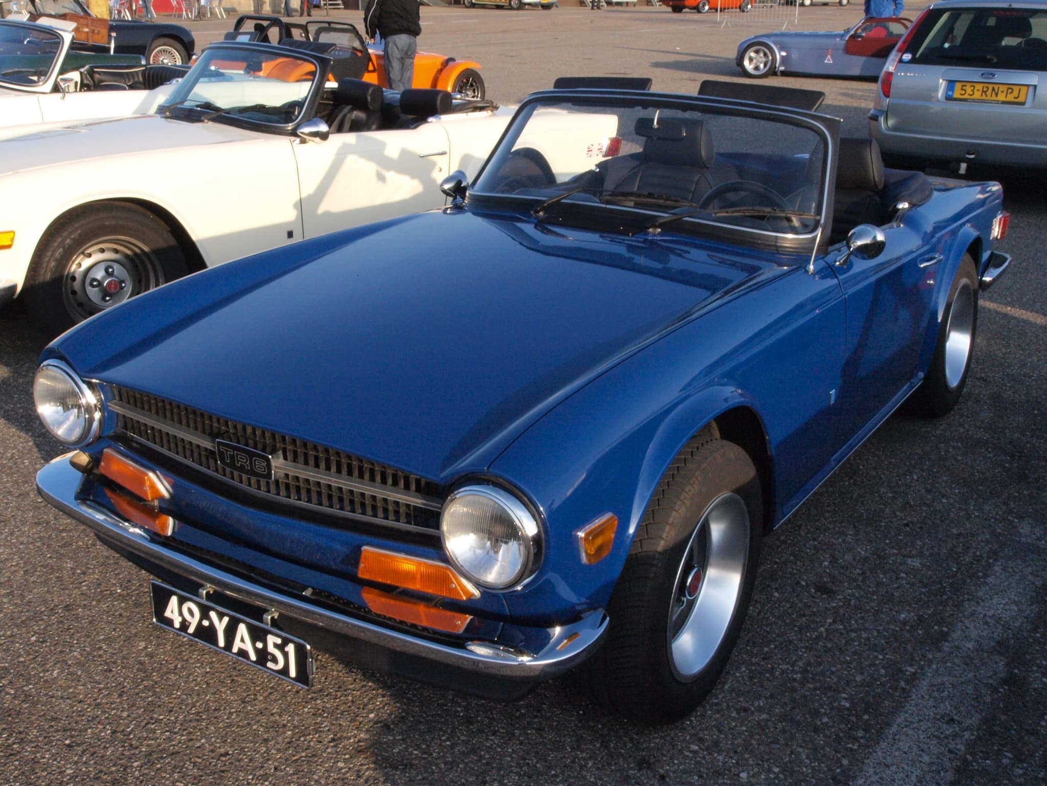 A dark blue Triumph TR6 with the hood folded in a paddock car park, in low evening sun.
