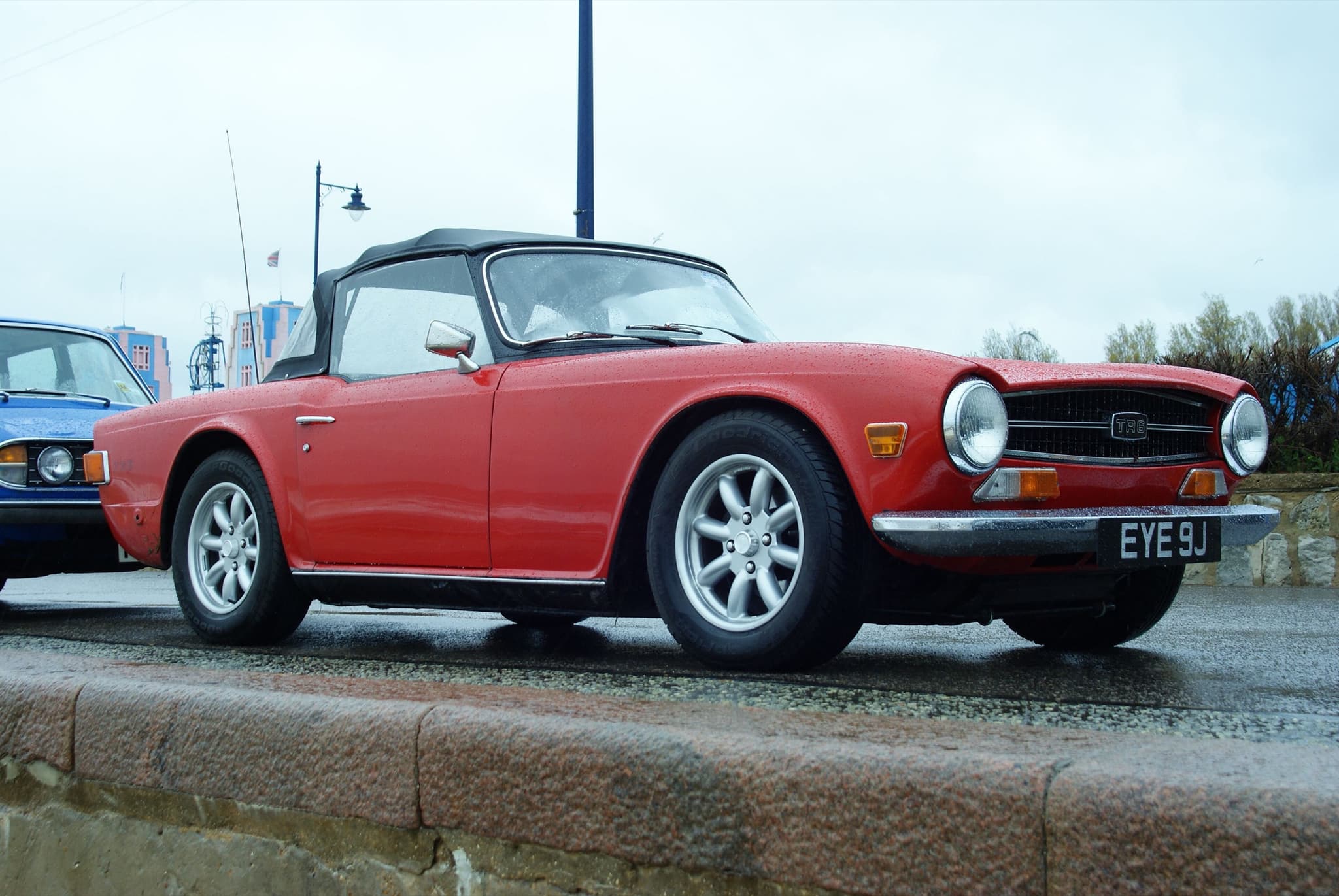 A red Triumph TR6 with its black hood up, parked on a wet seafront promenade under a grey sky. Opens a larger view
