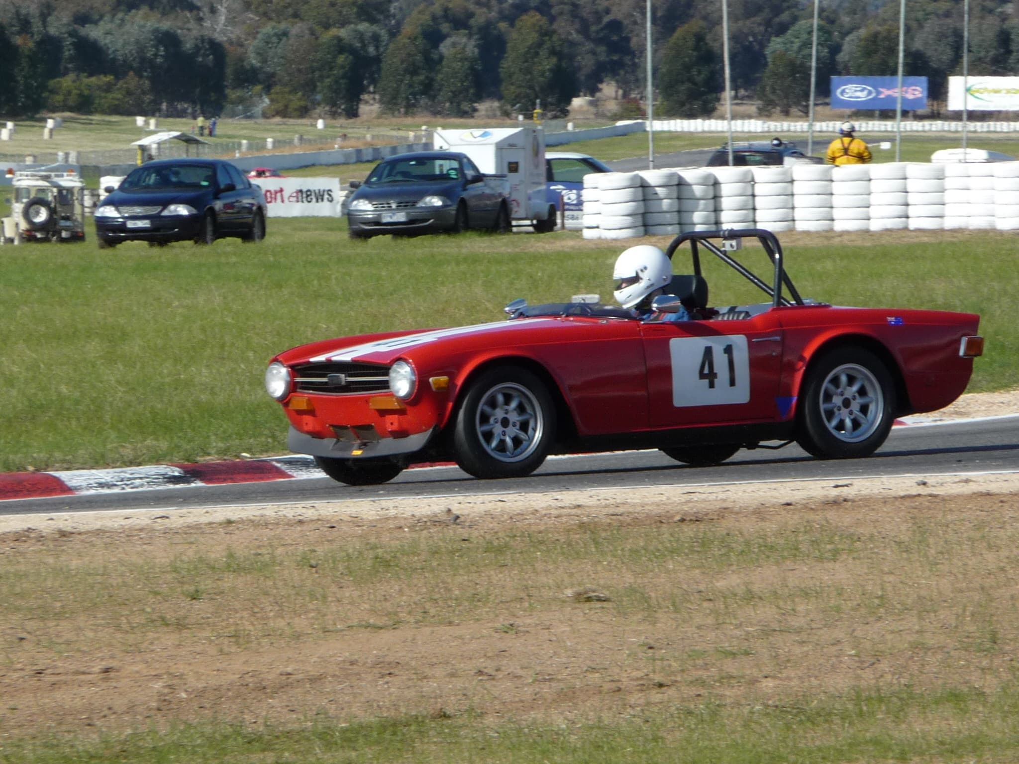 A red Triumph TR6 race car, number 41, with a roll cage and white bonnet stripes, on a circuit kerb with a tyre wall behind.