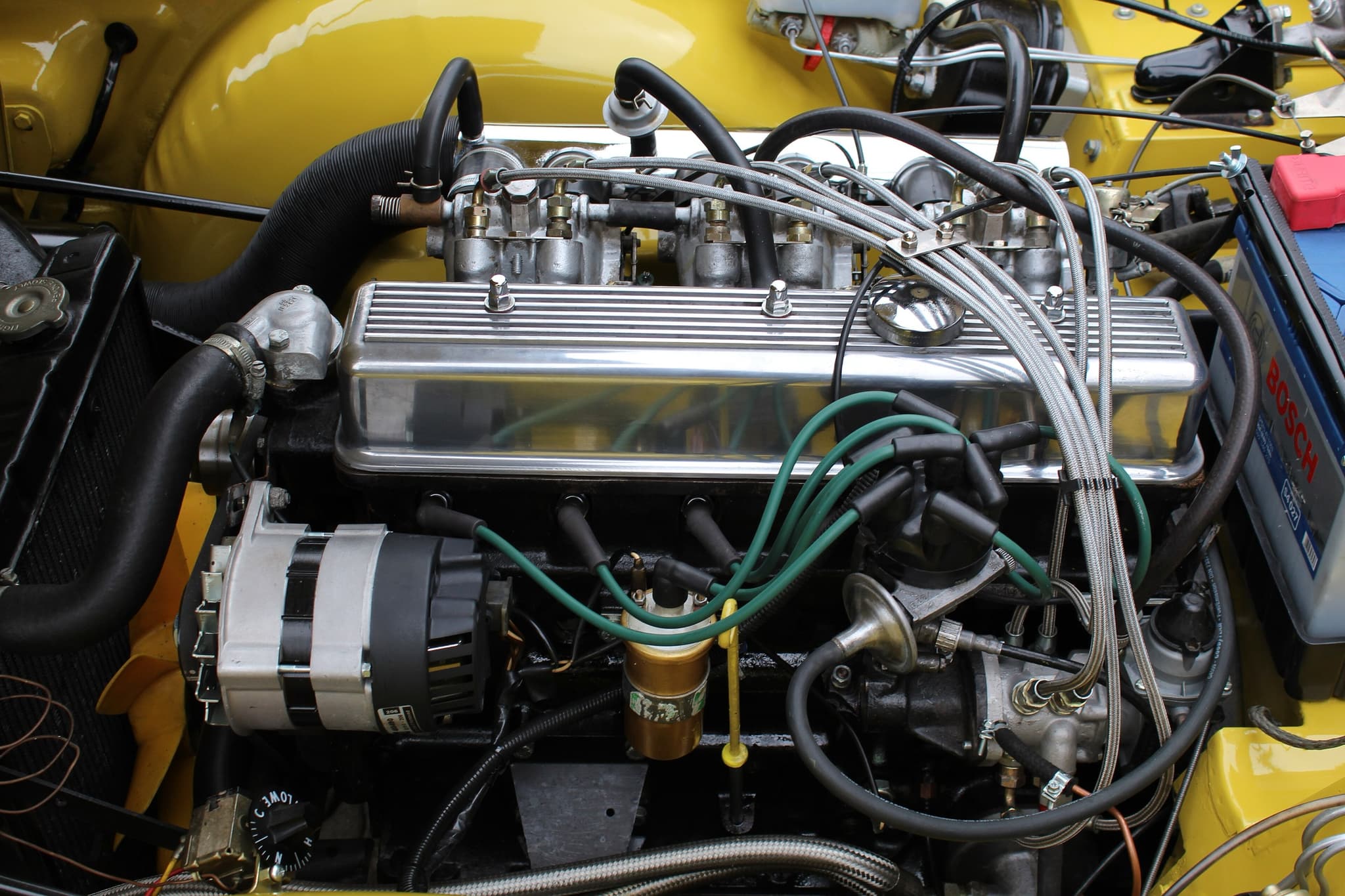 The petrol-injection six of a yellow Triumph TR6 PI: paired throttle bodies with brass injectors, braided injector pipes from the metering unit and a polished finned rocker cover.
