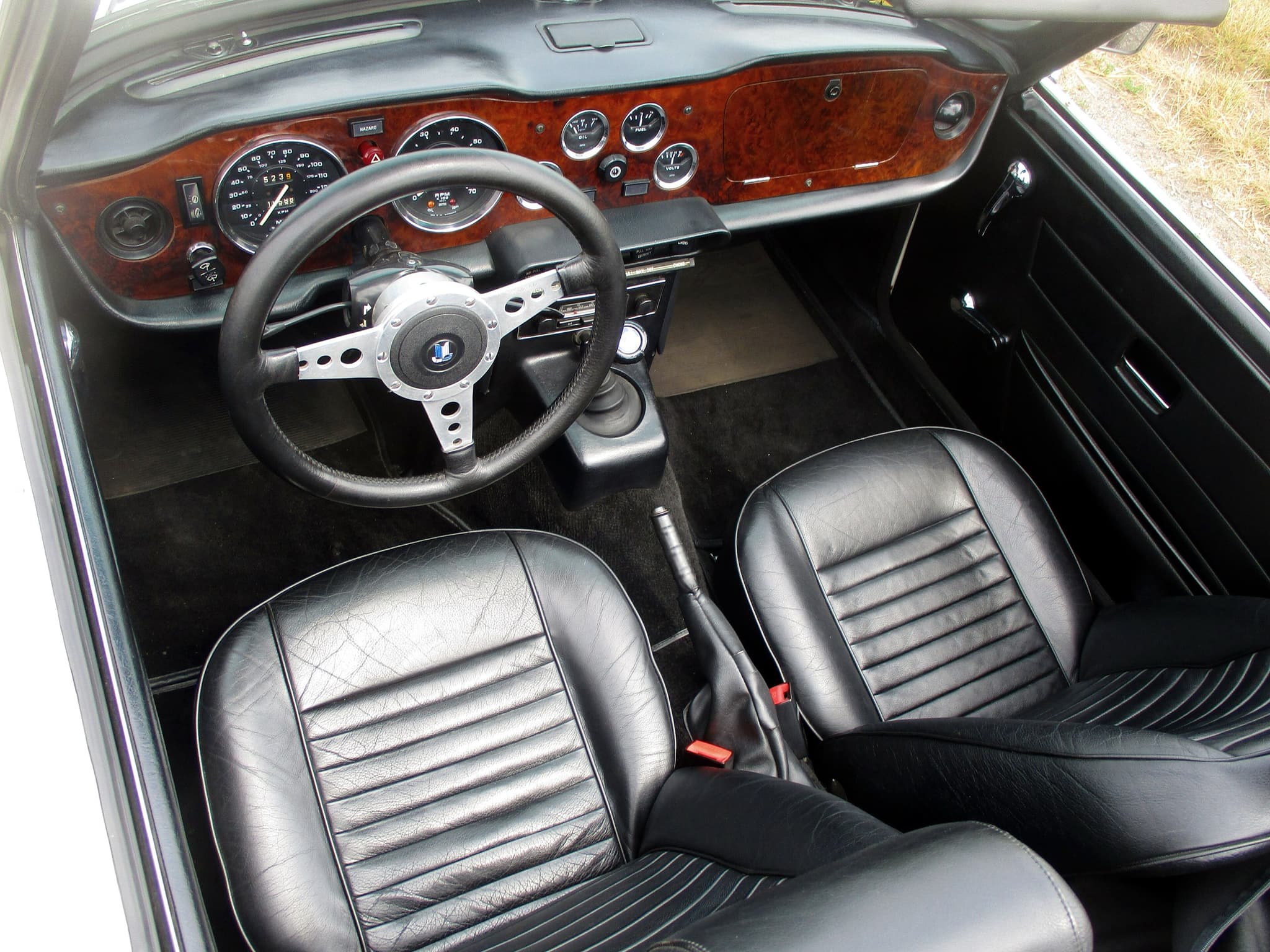 Looking down into a restored Triumph TR6 cockpit with retrimmed black leather seats piped in white, a wood facia with the later small gauges and a three-spoke steering wheel.