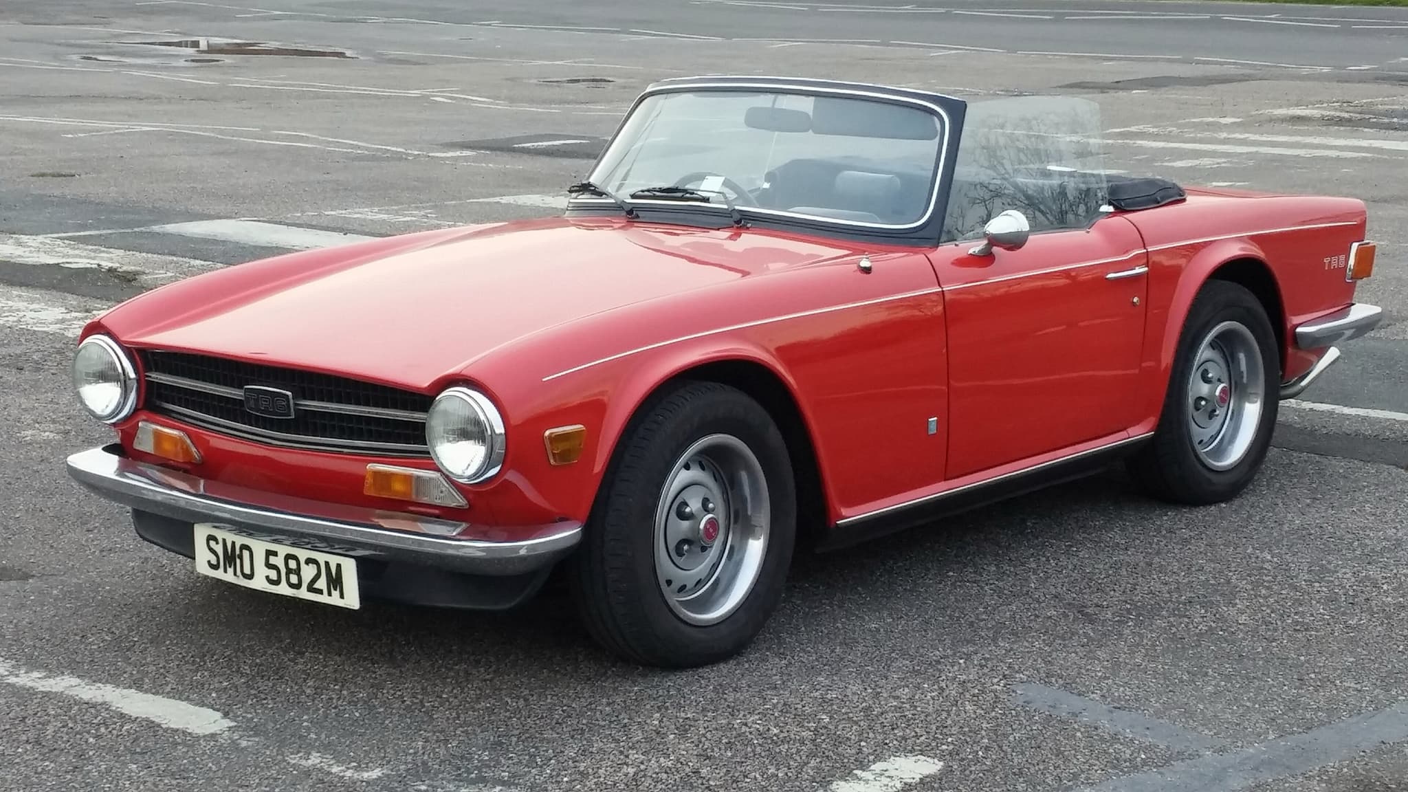 A red right-hand-drive Triumph TR6 with its hood folded and a black front spoiler, front three-quarter in an empty car park. Opens a larger view