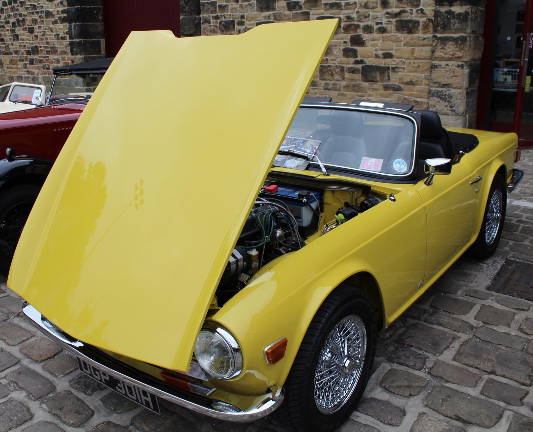 A yellow Triumph TR6 with its front-hinged bonnet raised to show the engine, on wire wheels before a stone building.