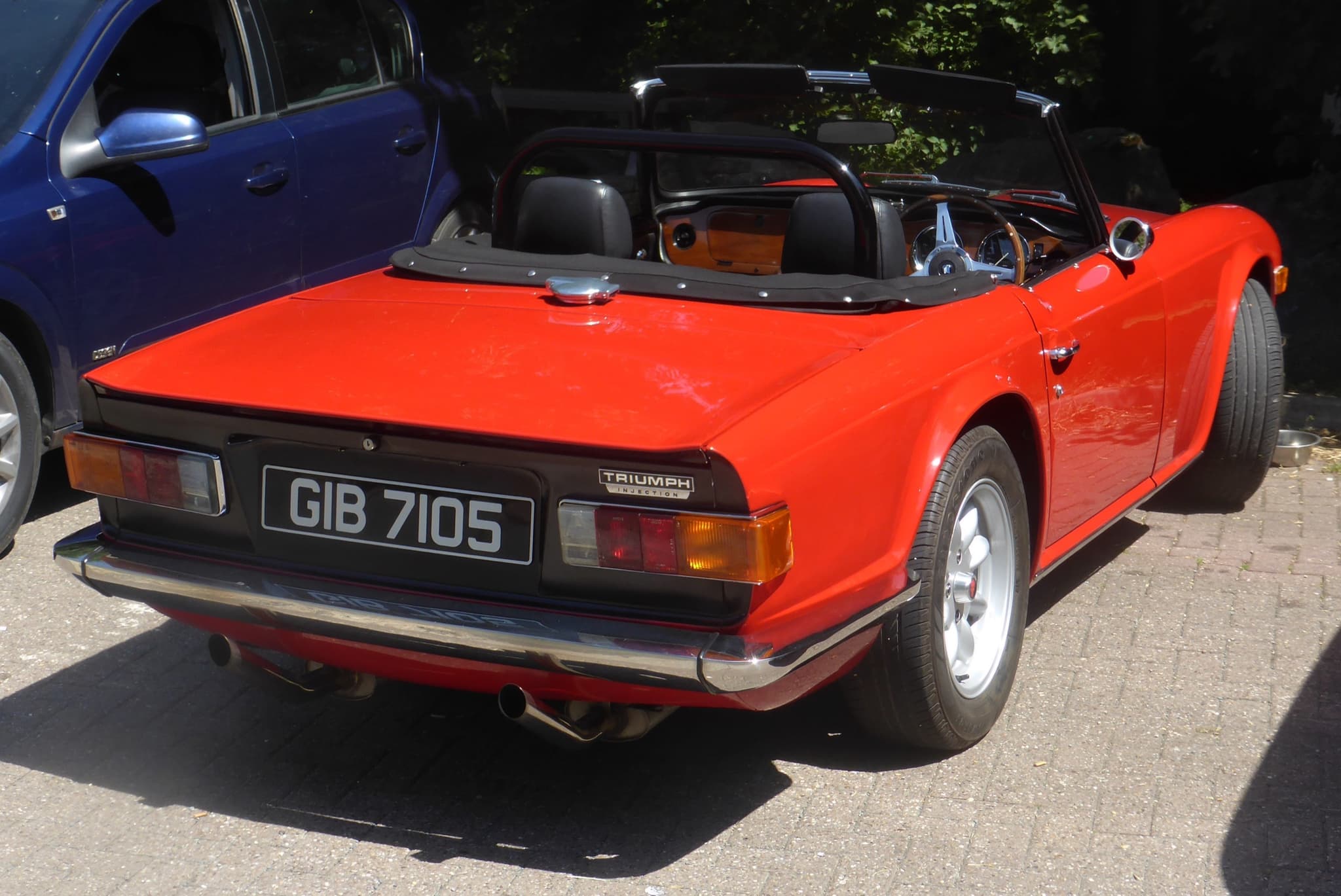 A red Triumph TR6 seen from behind on block paving, its TRIUMPH INJECTION badge on the black rear panel. Opens a larger view