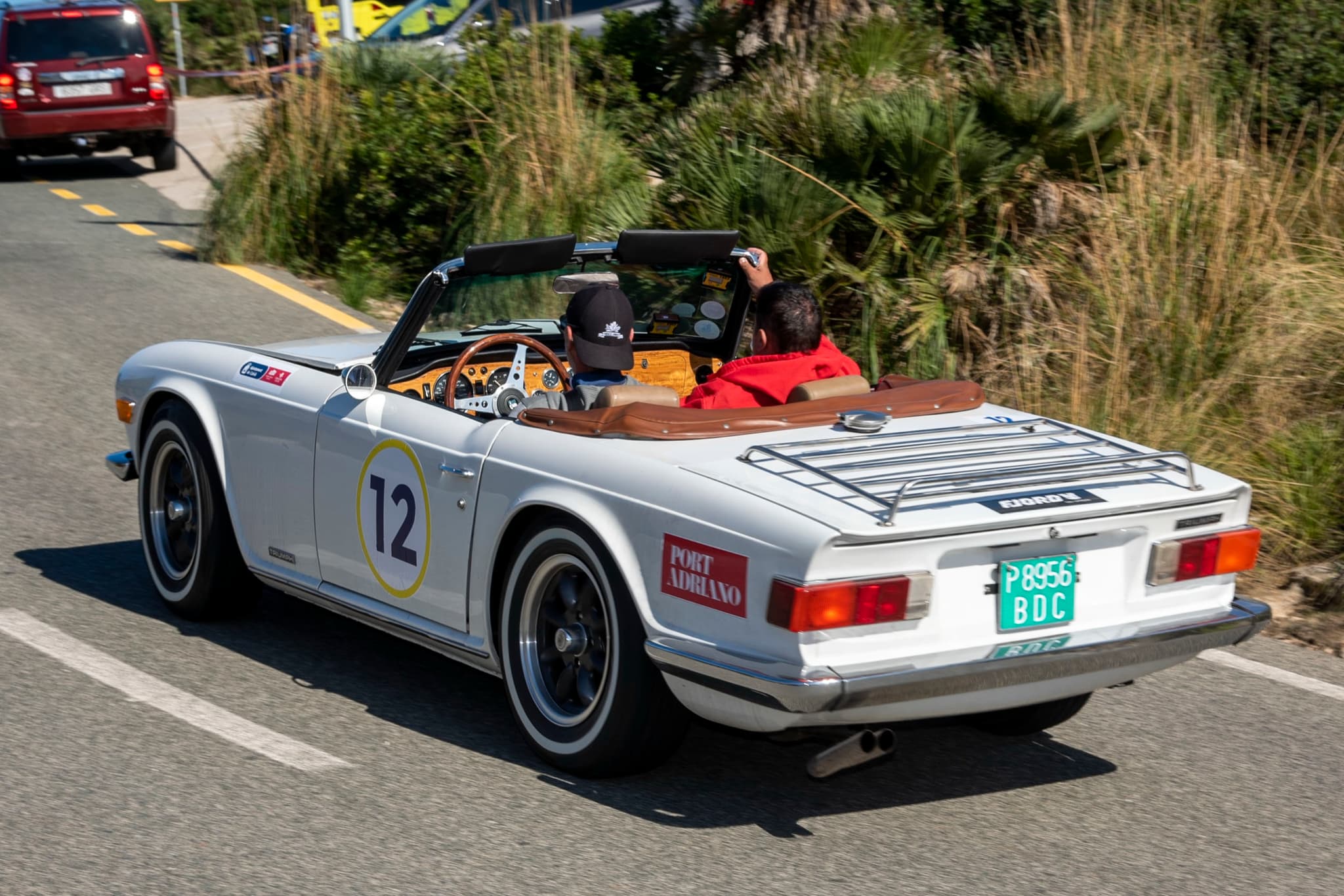 A white Triumph TR6 with start number 12 and a luggage rack, seen from behind on a mountain road during a classic rally.