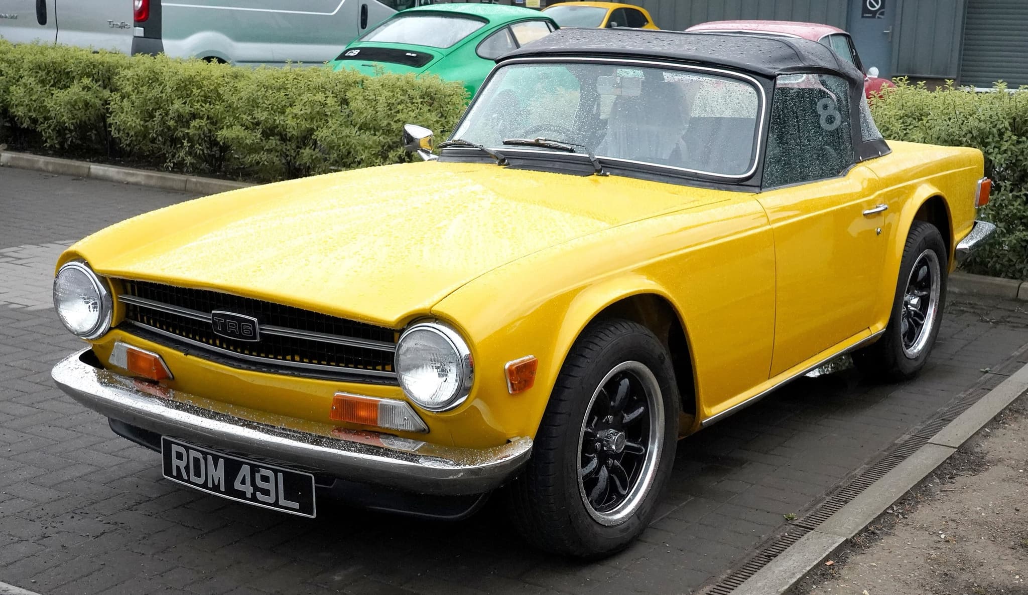A yellow Triumph TR6 with its black hood up, parked in the rain on block paving outside a cafe.