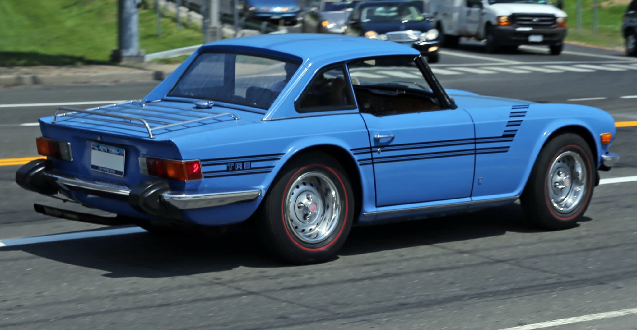 A blue Triumph TR6 with a matching hardtop and a luggage rack, driving away along a city road.
