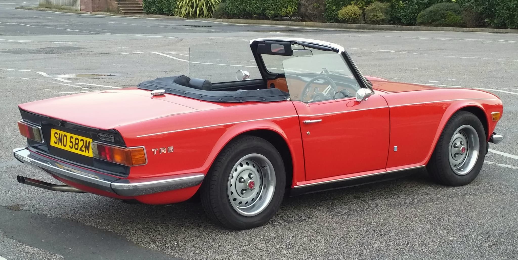A red right-hand-drive Triumph TR6 seen from behind in an empty car park, showing its black rear panel and wrap-round tail lamps.