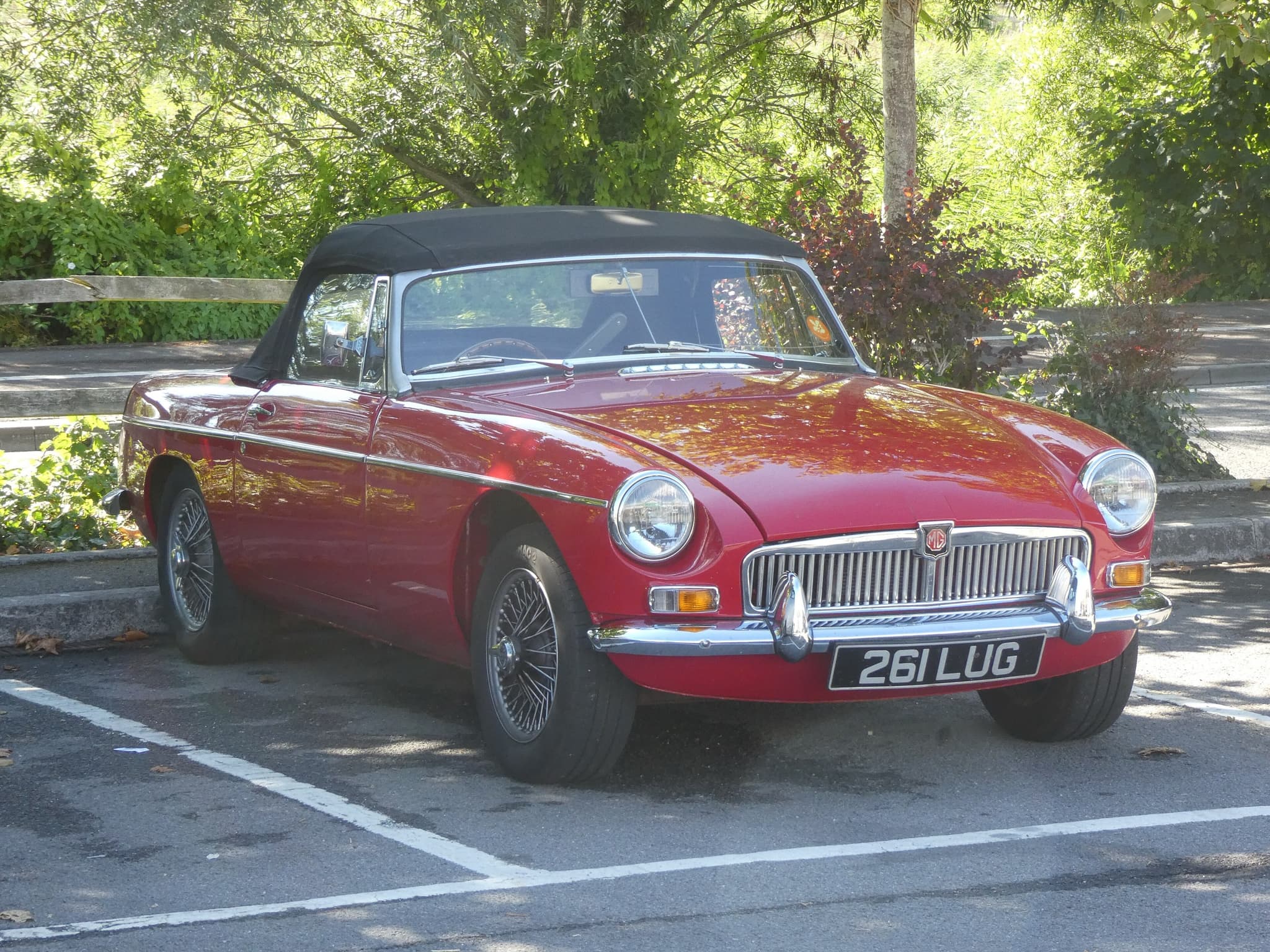 A red sports car with a black hood up, chrome bumpers, a vertical-slat grille and wire wheels, parked in a car park under trees.